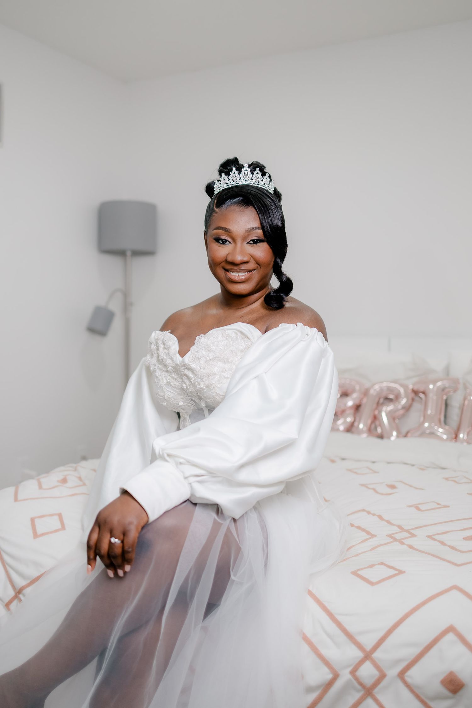 A bride in a white off-shoulder wedding dress poses on a bed with geometric patterned bedding in a bright room.