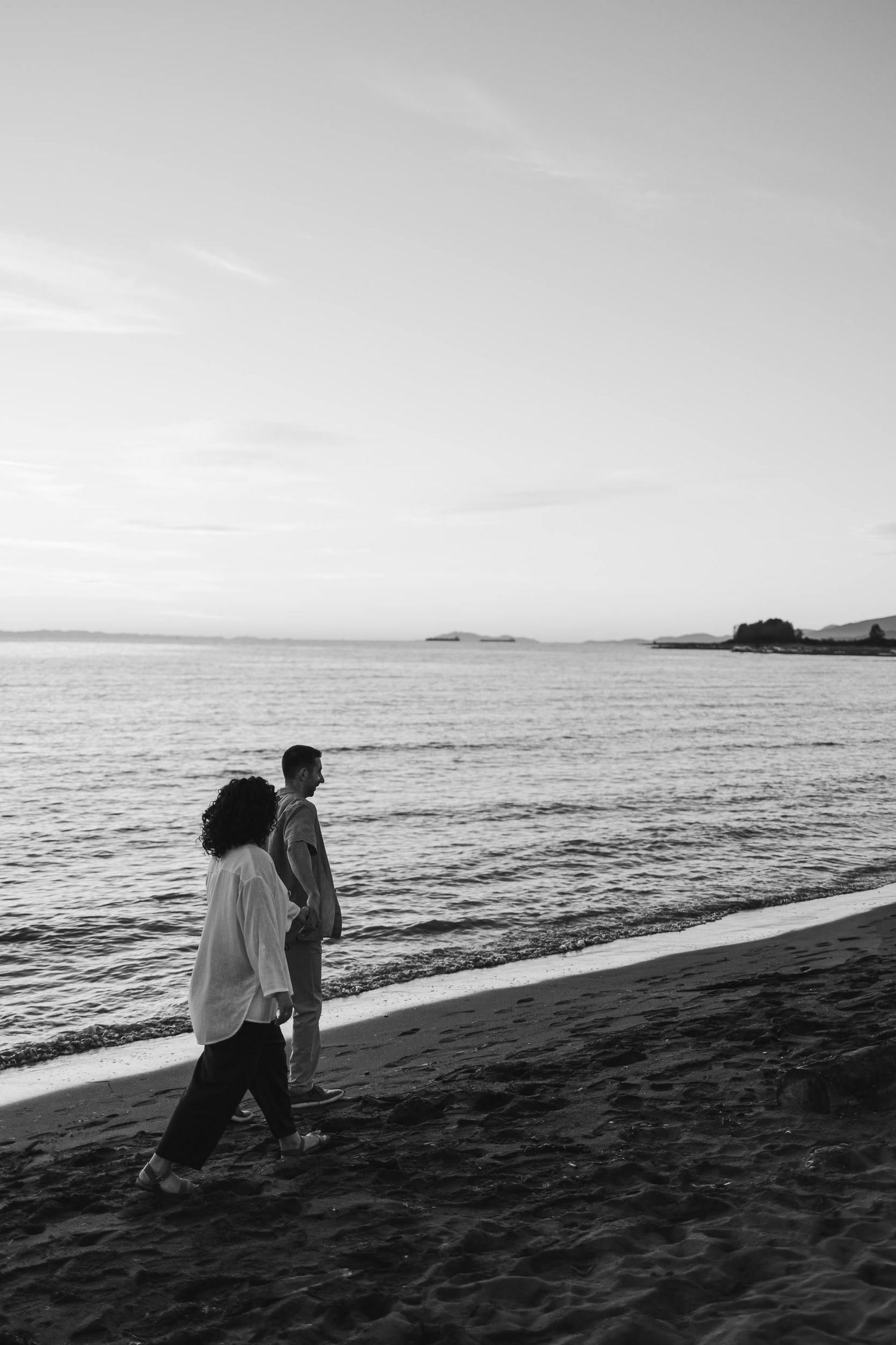 Two silhouettes walk along a black sand beach at sunset with ocean waves and mountains in the distance.