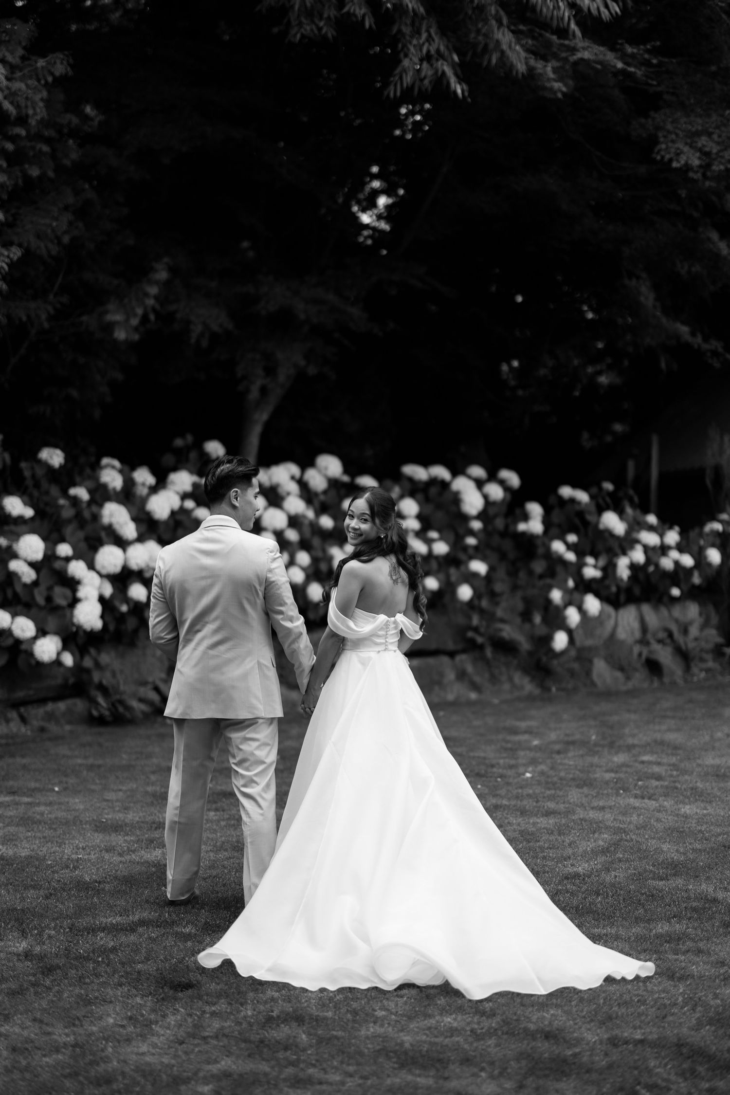 Black and white photo of bride and groom walking hand in hand through a lush garden with blooming hydrangeas.