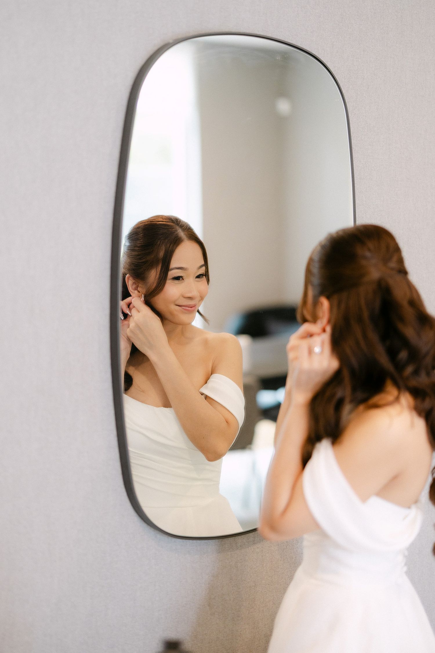 Bride in white off-shoulder dress putting on earrings while looking at her reflection in a modern mirror.