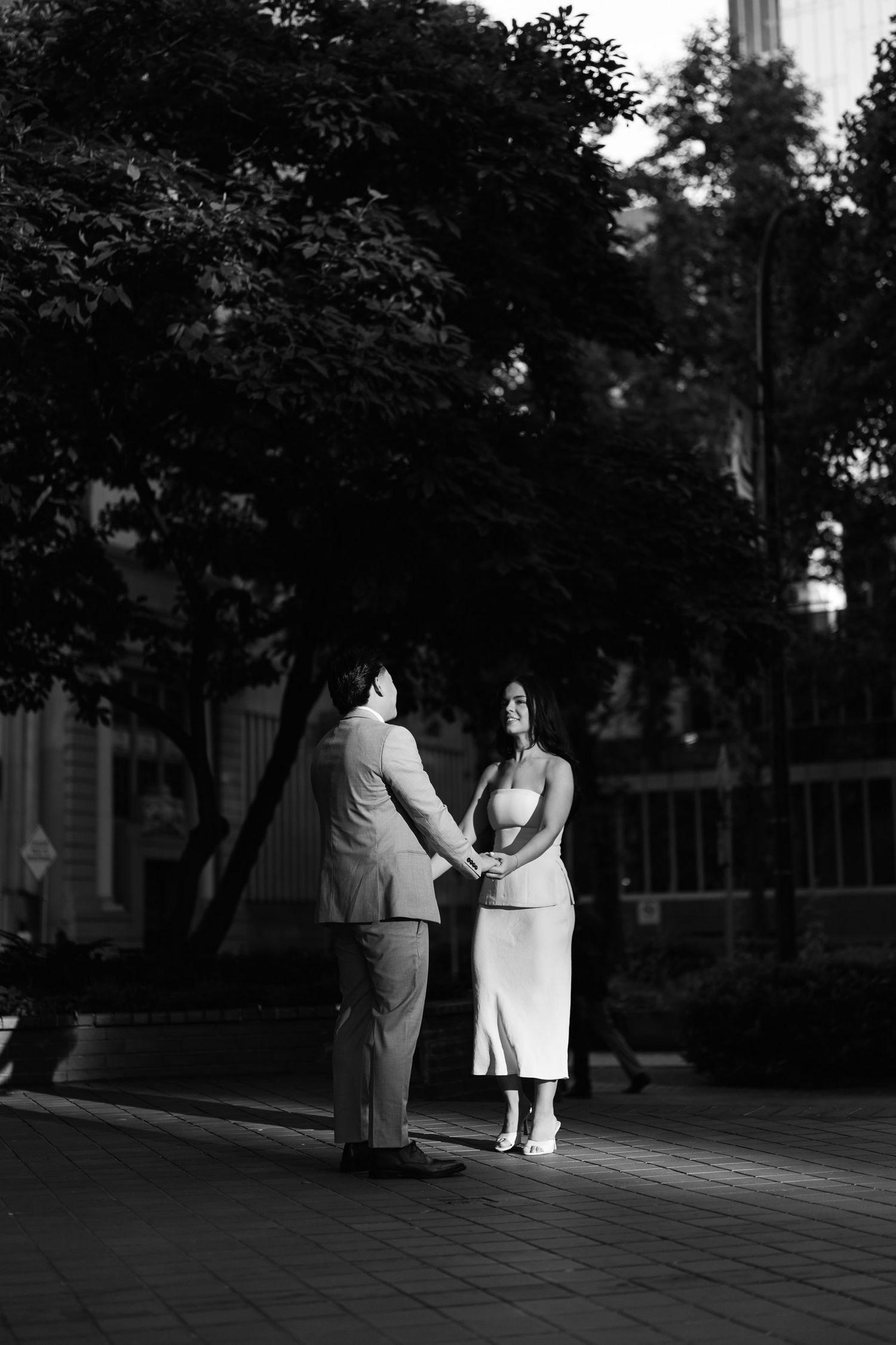 Black and white photo of an engaged couple holding hands beneath large trees in a downtown plaza.