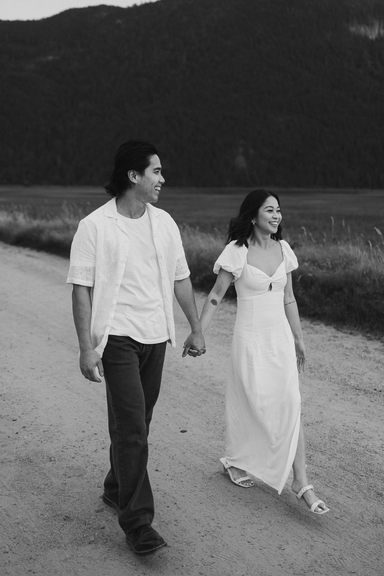 Black and white photo of a smiling couple holding hands and walking down a dirt road with a forested hillside behind them.