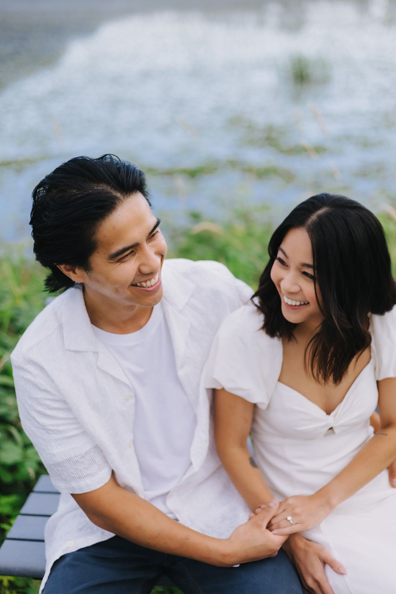 Smiling couple in white outfits sit close on a bench holding hands beside a sparkling lake, a joyful engagement or couples portrait.