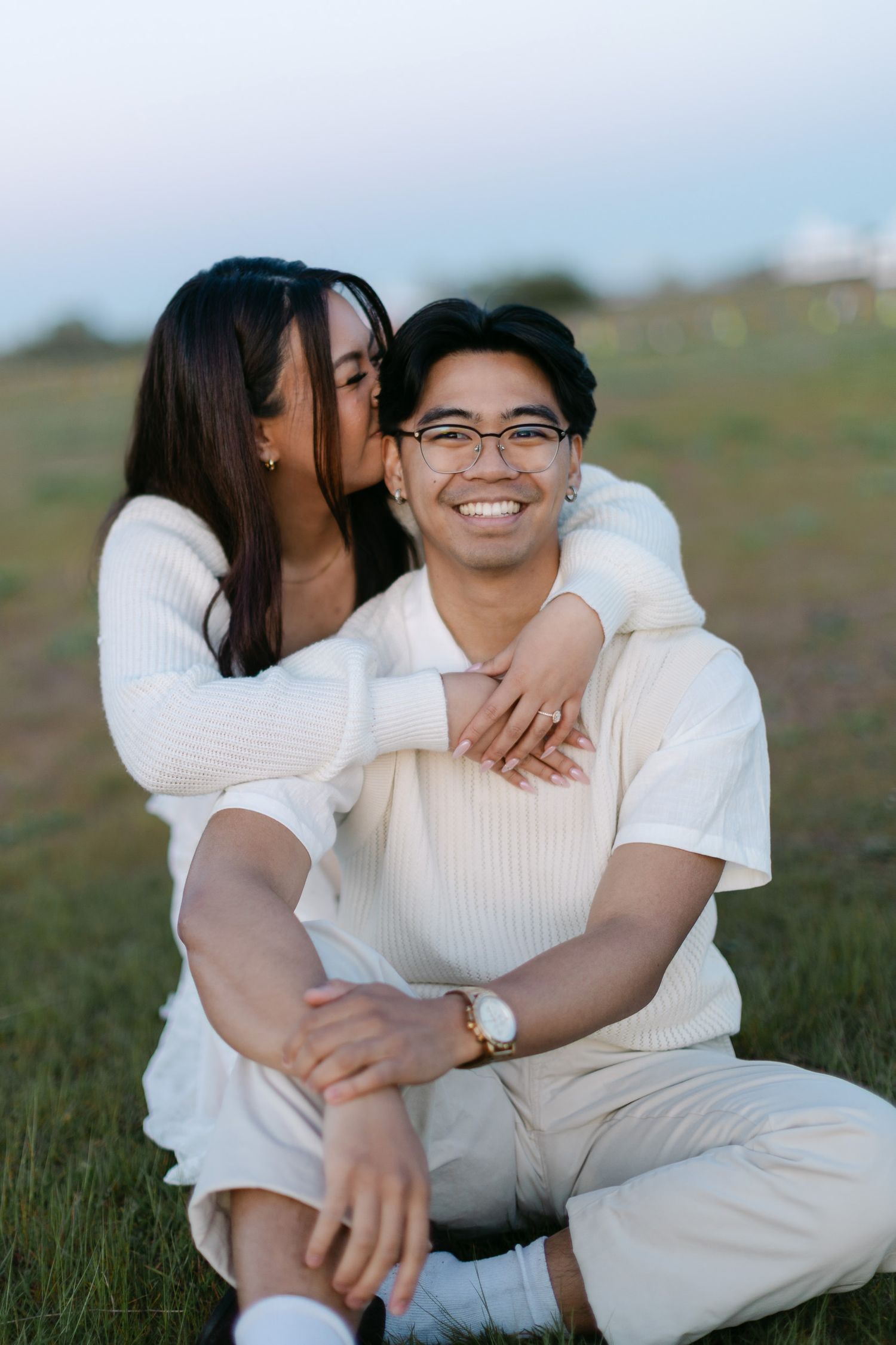 Smiling man in glasses sits in a grassy field as a woman hugs him from behind and kisses his cheek, both in cream outfits.