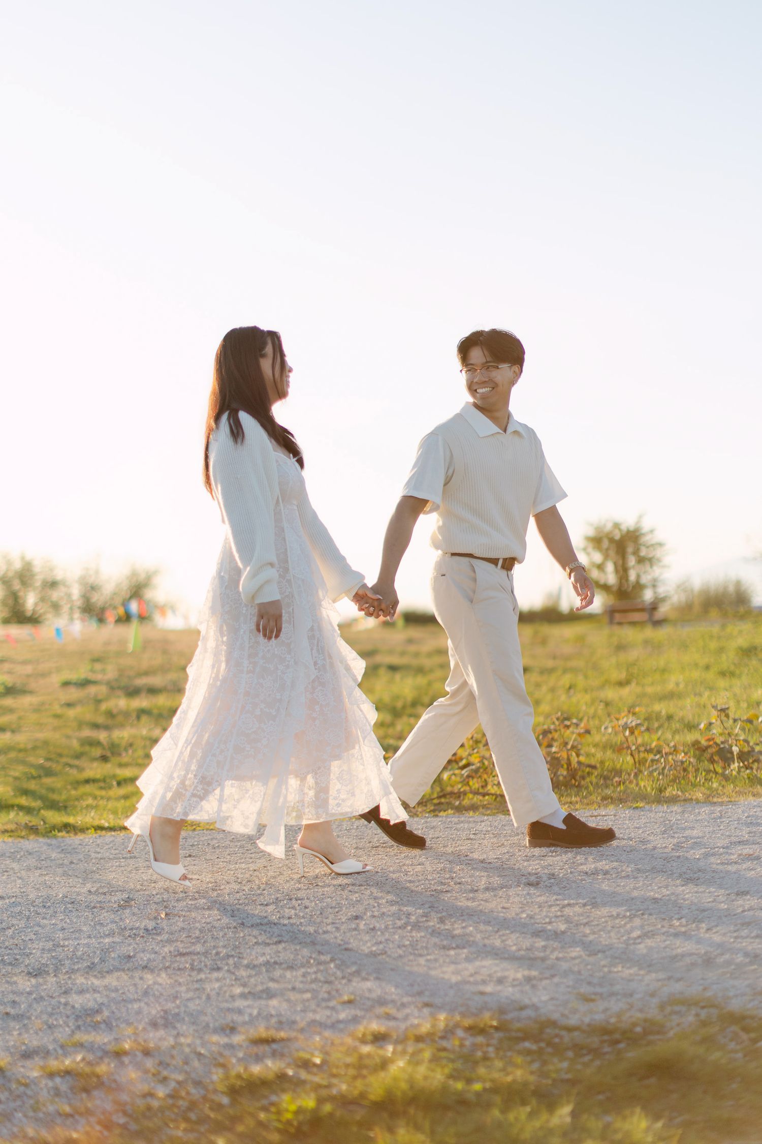 Smiling couple in white outfits holding hands and walking along a gravel path through a sunny grassy field at golden hour.