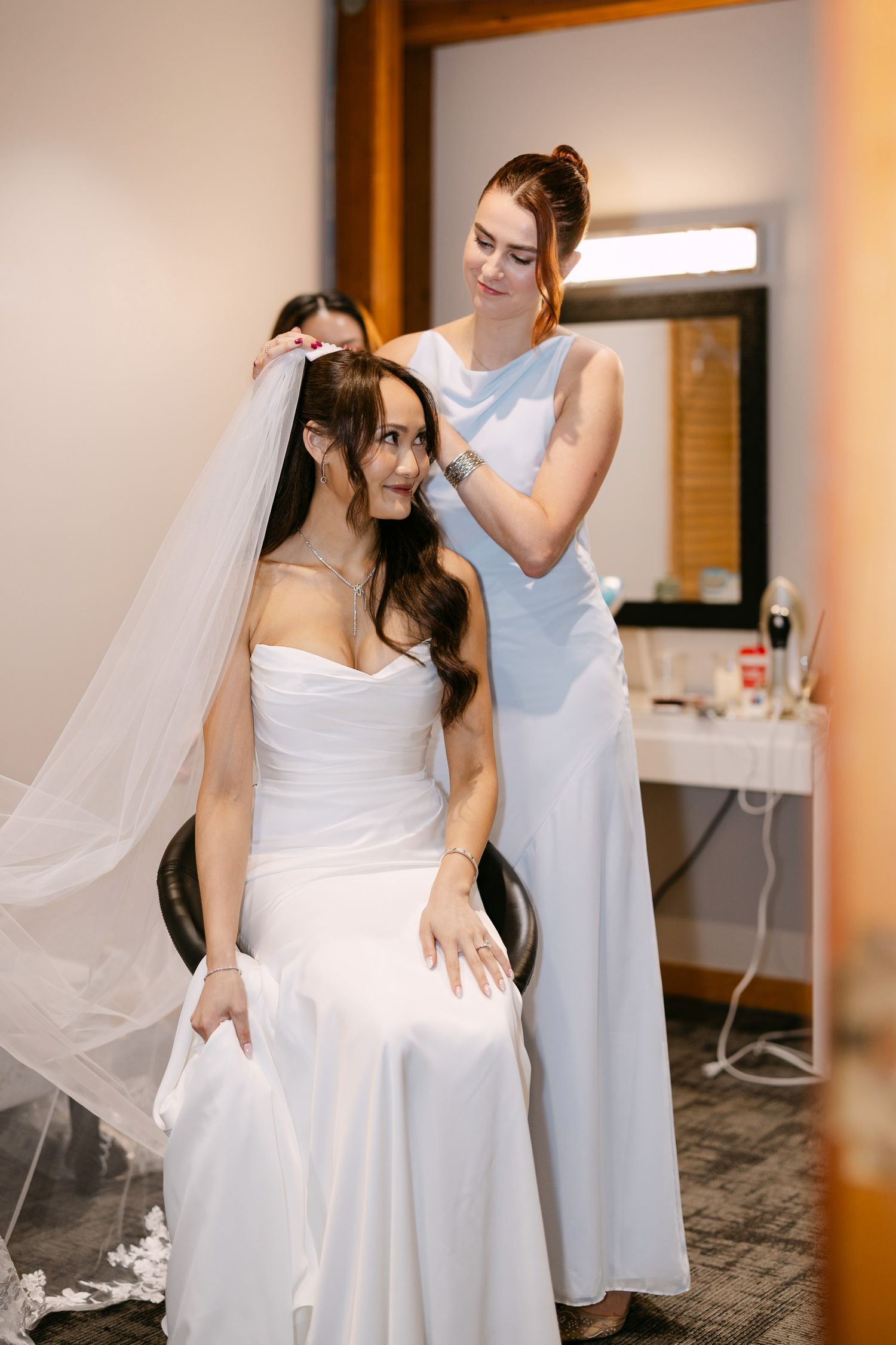 Bridesmaid in light blue dress adjusting veil on smiling bride in white strapless gown seated before mirror.