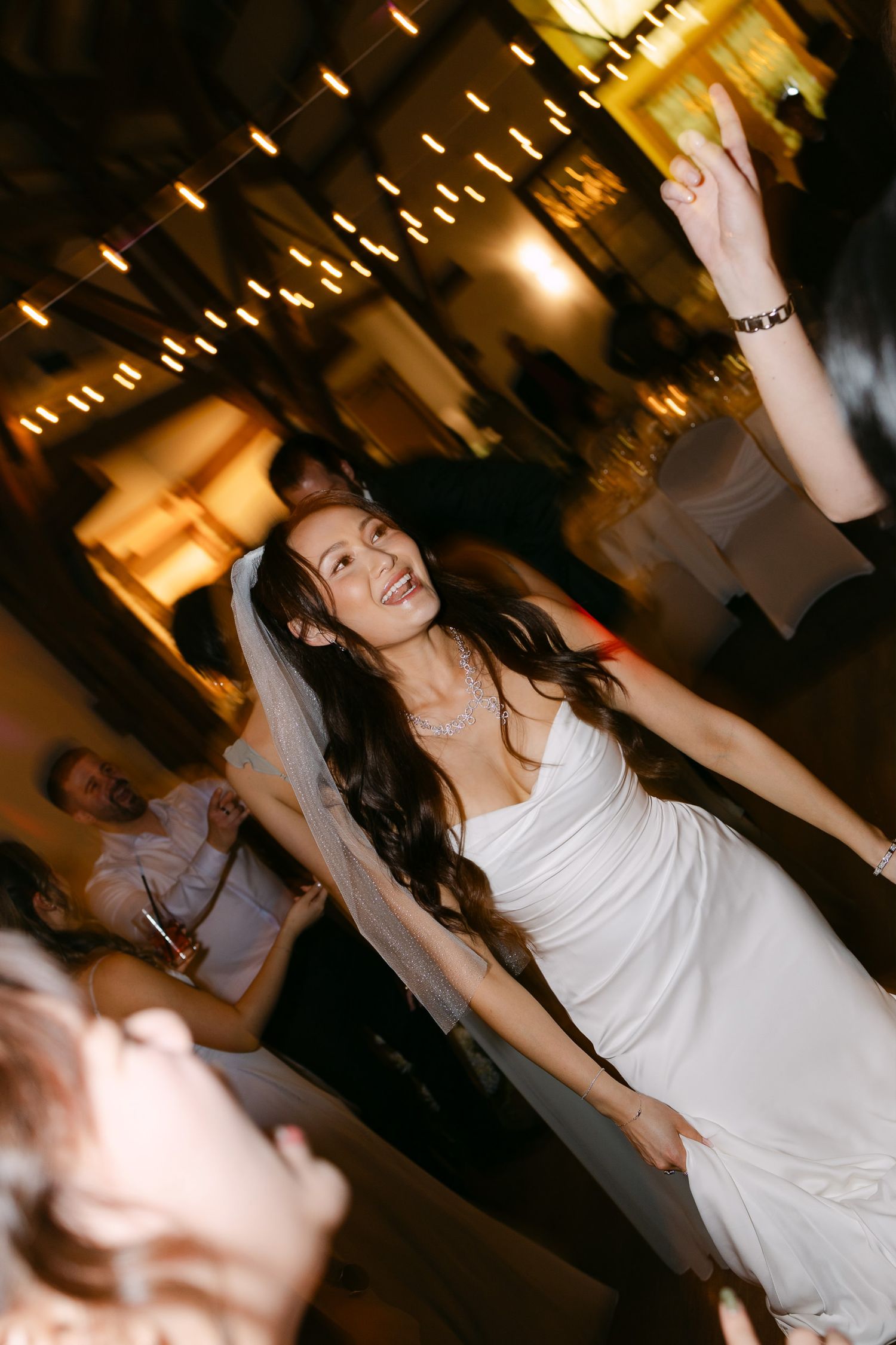 Bride in white dress dancing joyfully at wedding reception with string lights glowing in background.
