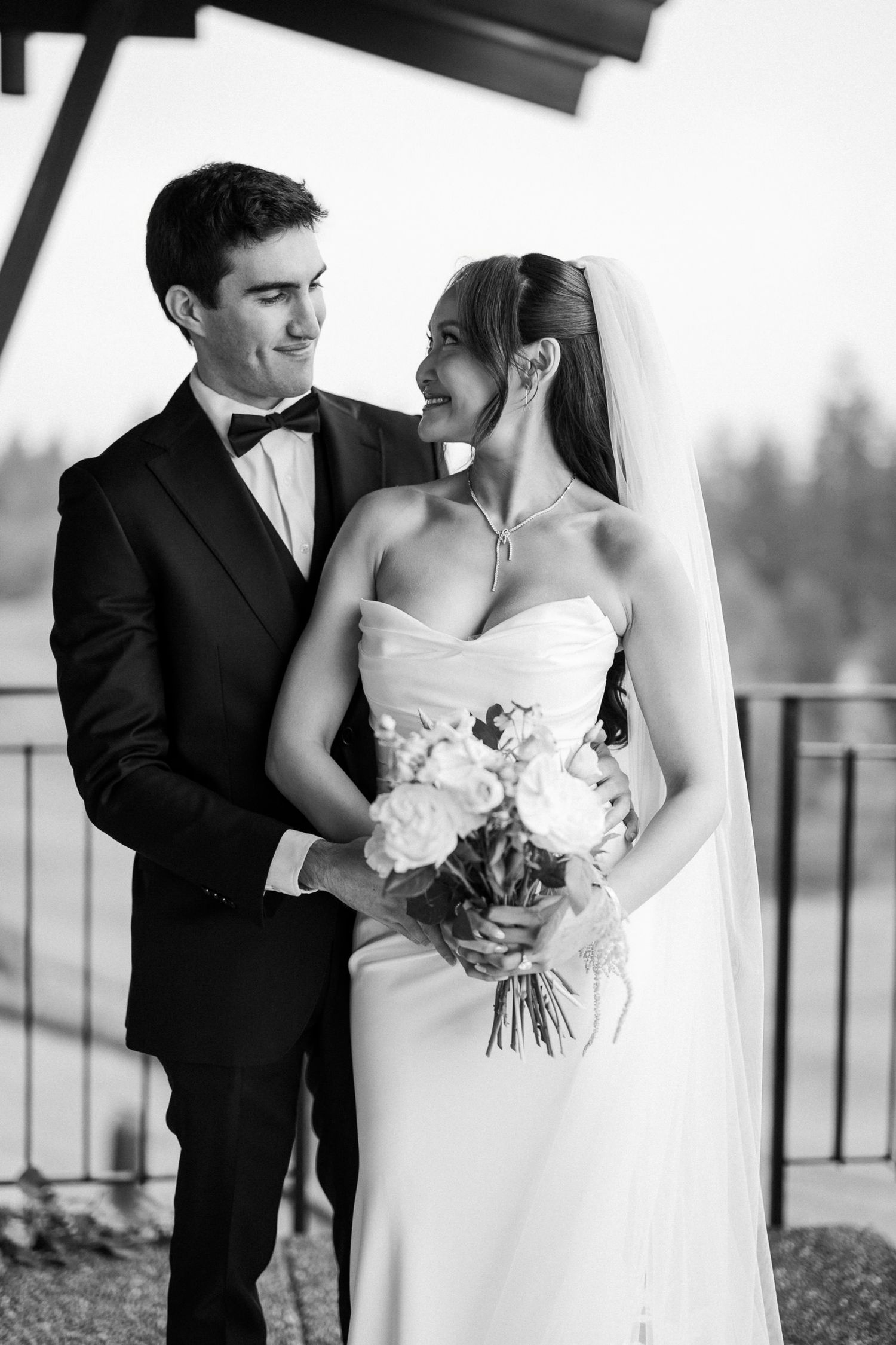Black and white photo of a smiling bride in strapless gown holding bouquet with groom in tuxedo at wedding.