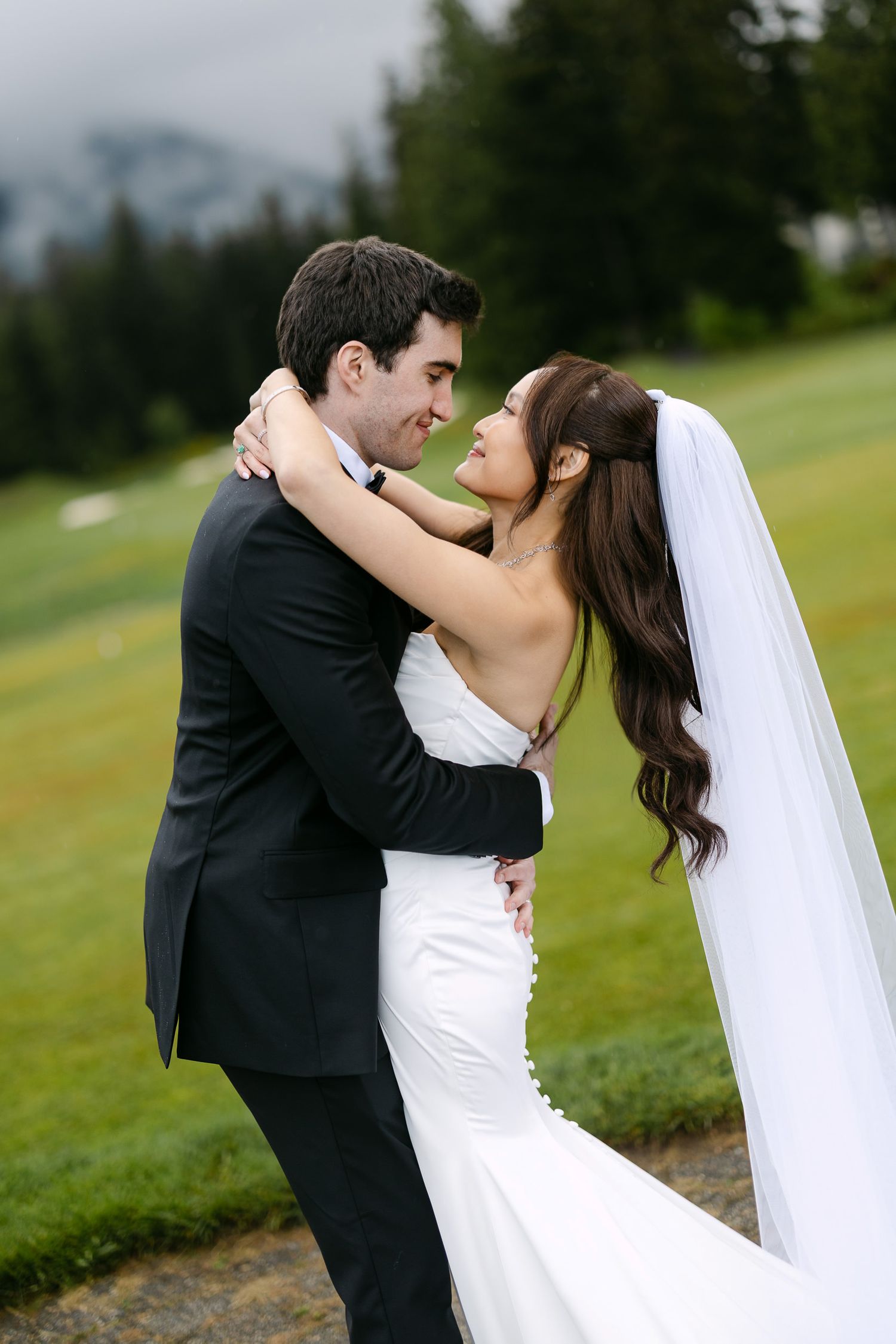 Bride and groom embrace on a lush green lawn with mountain backdrop on their wedding day.