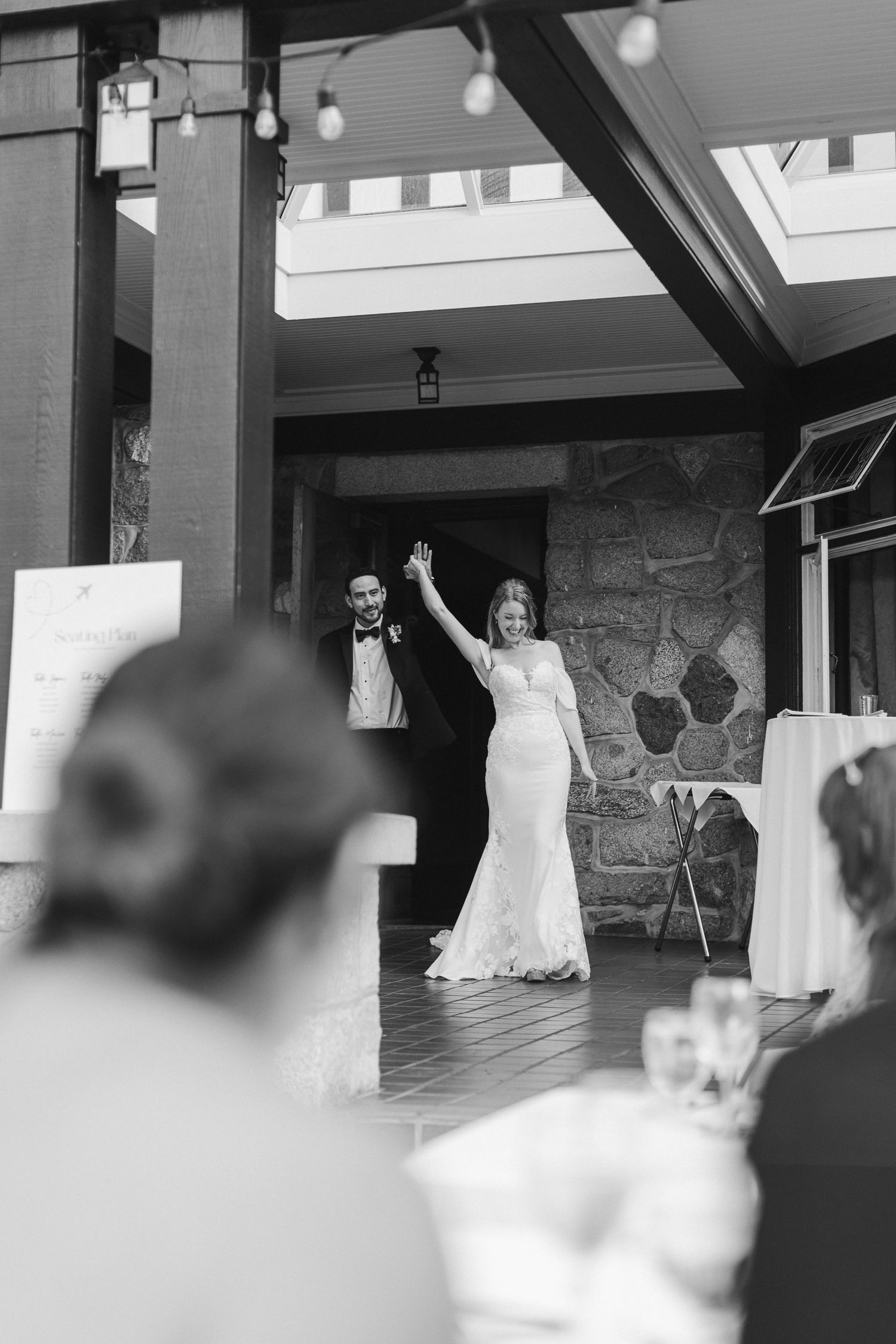 A black and white wedding celebration photo showing a couple raising their hands in triumph at an indoor-outdoor venue.