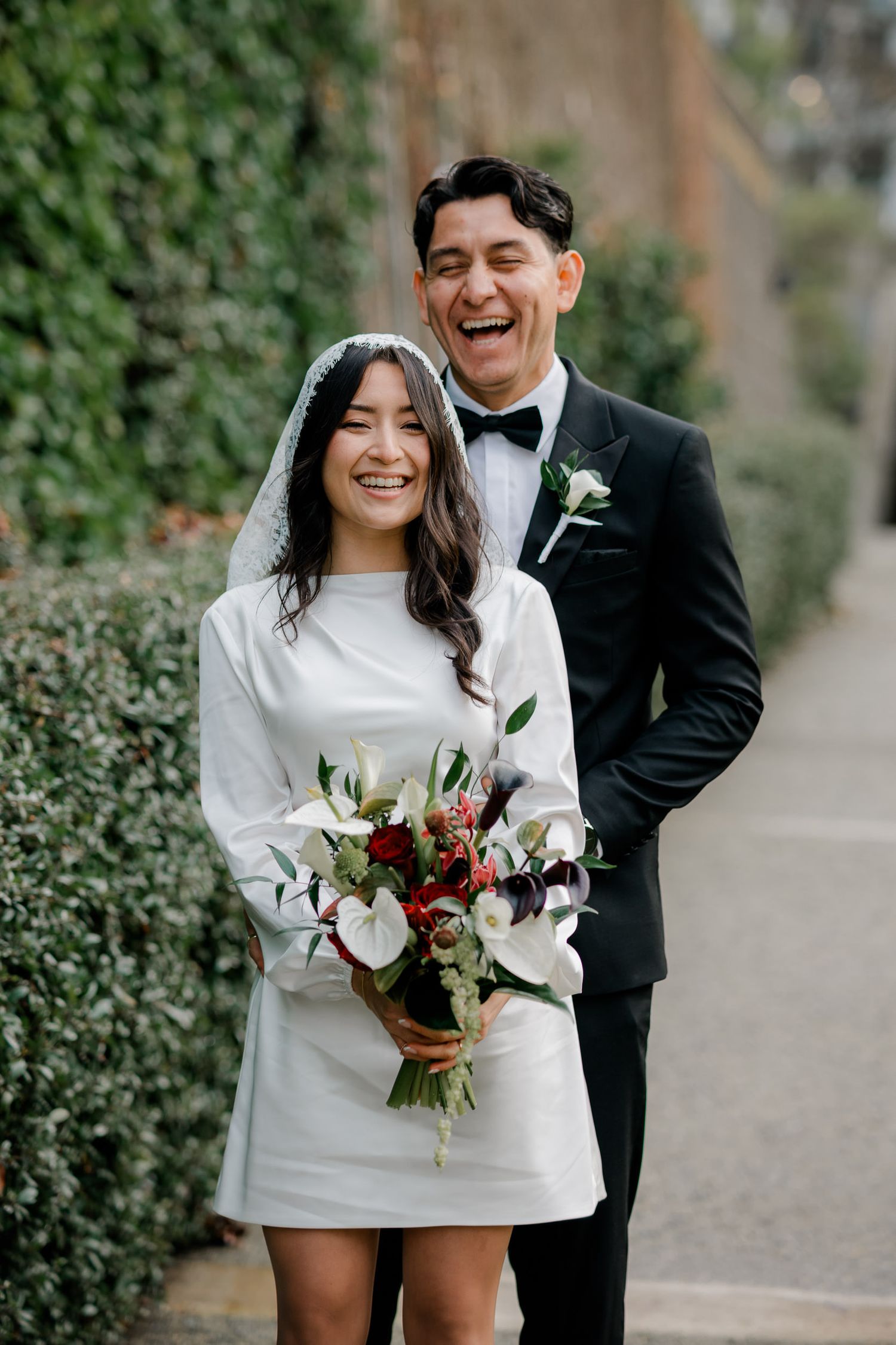 Joyful couple in wedding attire sharing candid moments together while holding a bouquet of red and white flowers.