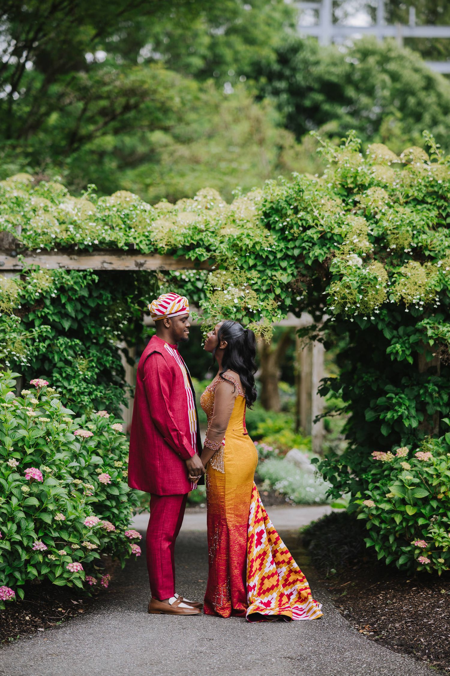 Two people in colorful attire walk together through a lush garden path lined with blooming flowers and greenery.