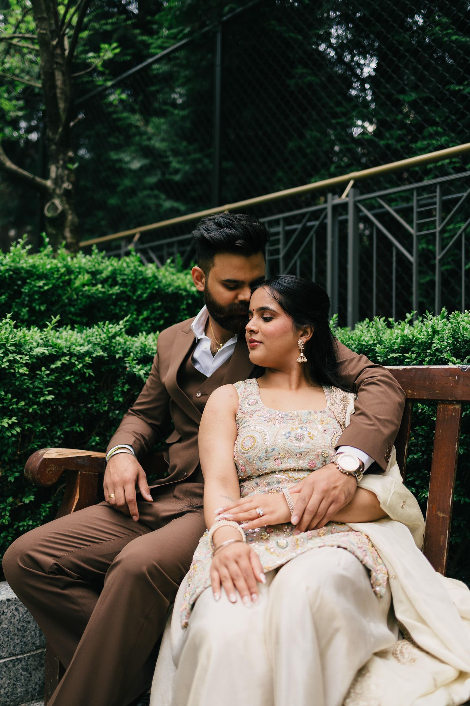 A romantic couple in formal attire share tender moments while sitting together on a wooden bench in a garden setting.