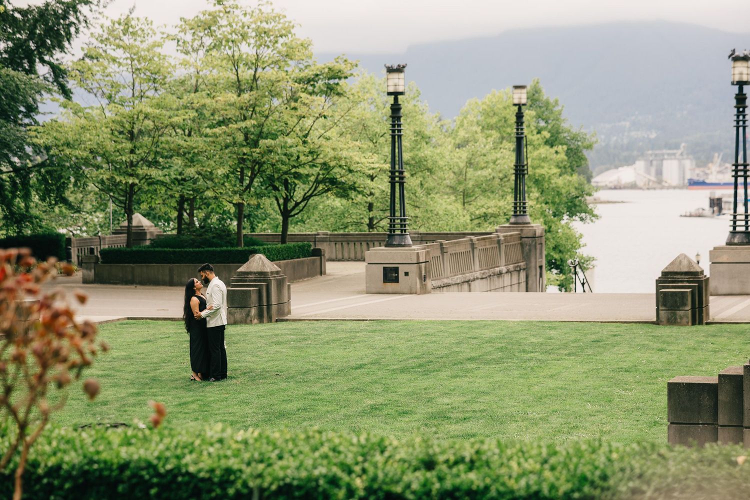 A panoramic view shows a lush green park with trees and pathways along a waterfront on a cloudy day.
