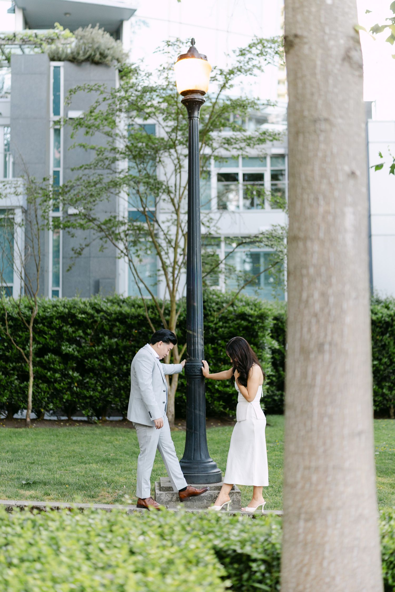 Couple in light summer attire pose beside a black lamppost on a green lawn with hedges and modern buildings in the background.