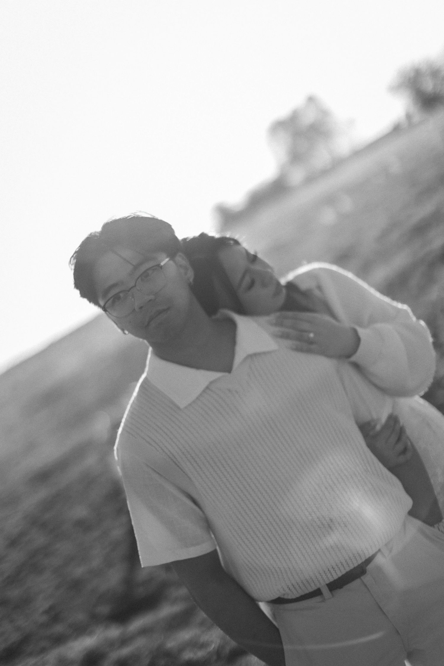 Black and white portrait of a man in glasses and knit vest as a woman rests her head on his shoulder from behind in a field.