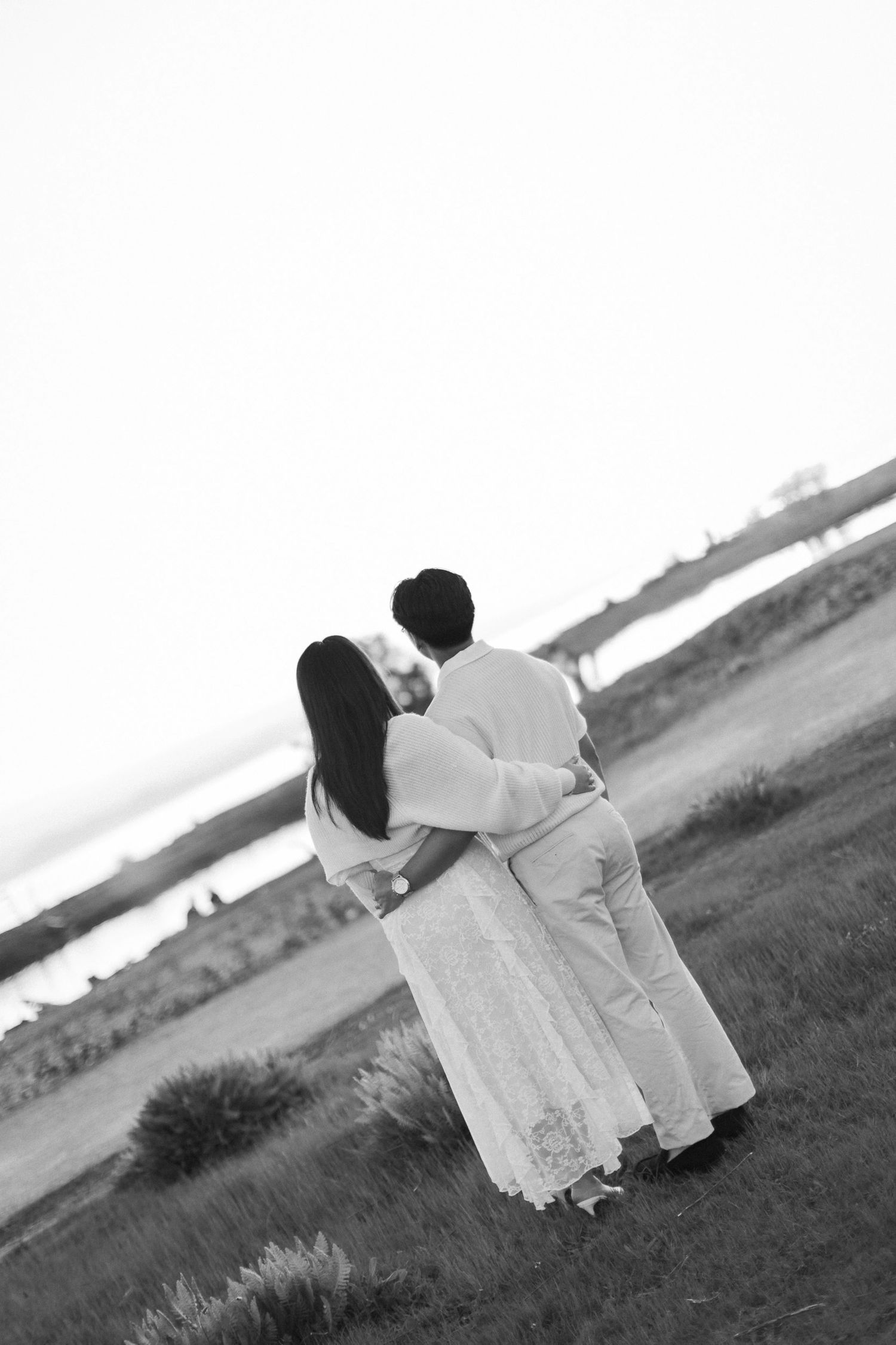 Black and white photo of a couple in white outfits embracing from behind while gazing at a calm waterfront from a grassy hill.