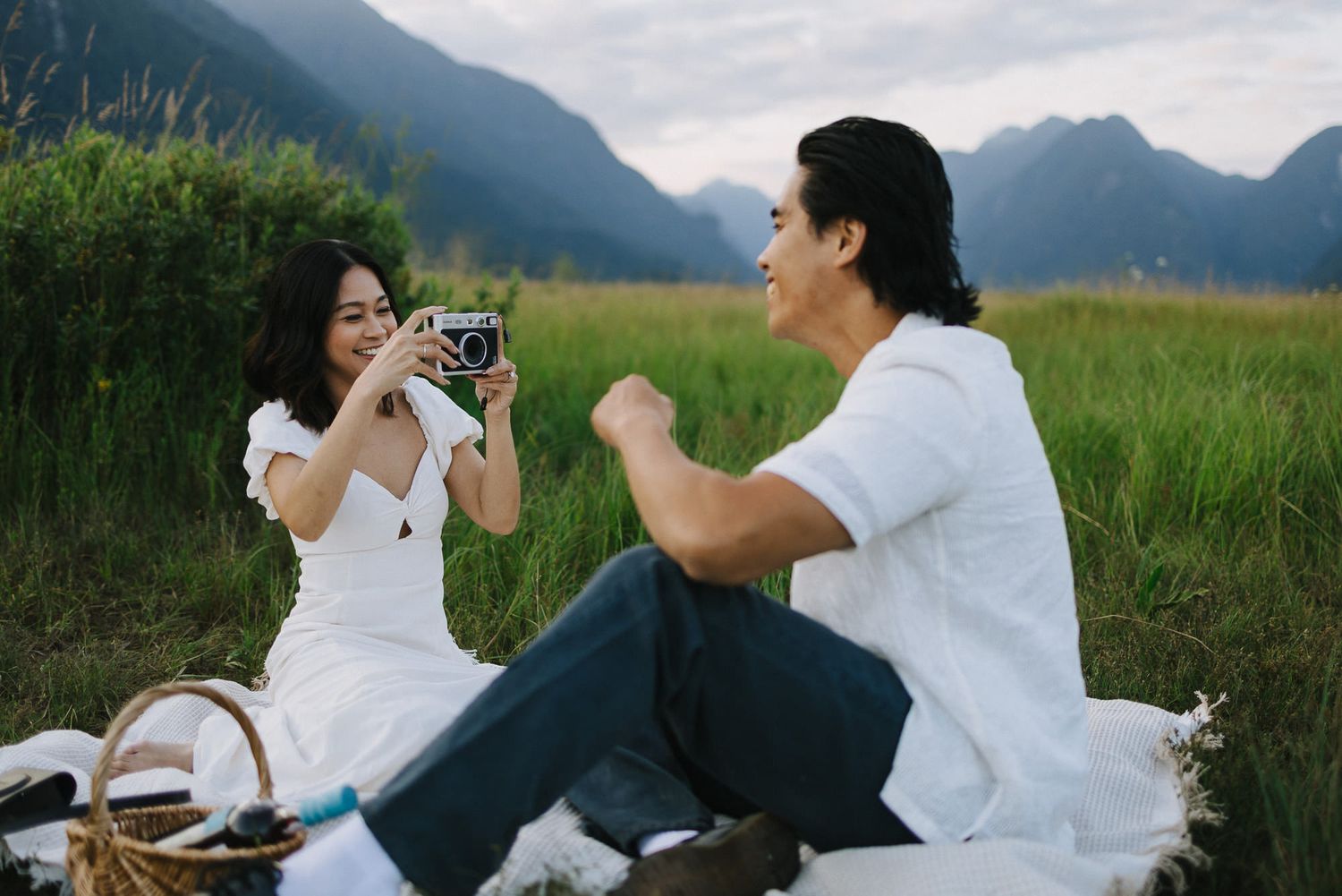 A woman in a white dress photographs her partner with a film camera during a picnic in a mountain meadow.