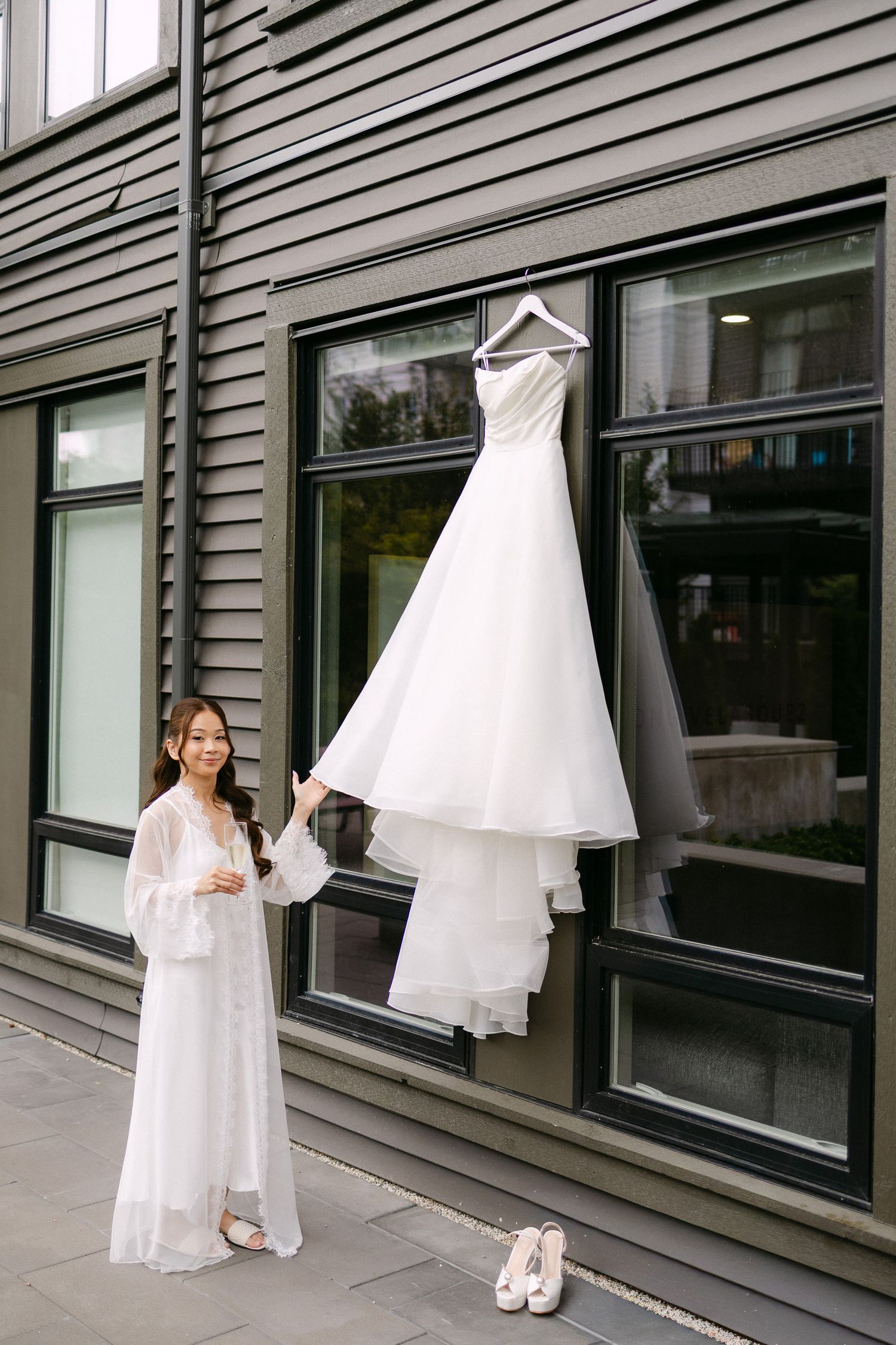 Bride in white robe stands beside her elegant wedding gown hanging on a hanger outside a modern building.