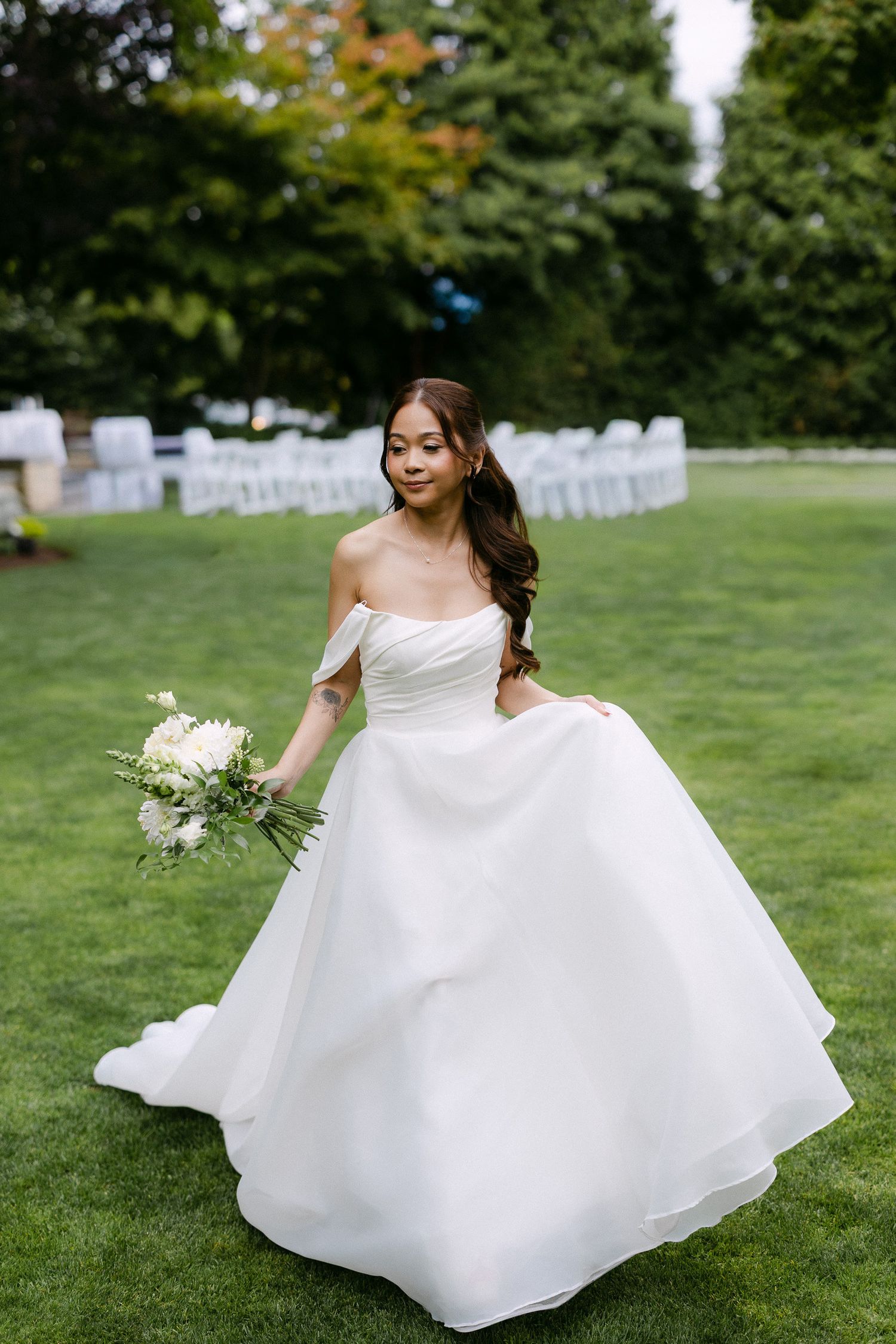 Bride in elegant off-shoulder white ballgown twirls on green lawn holding white floral bouquet outdoors.