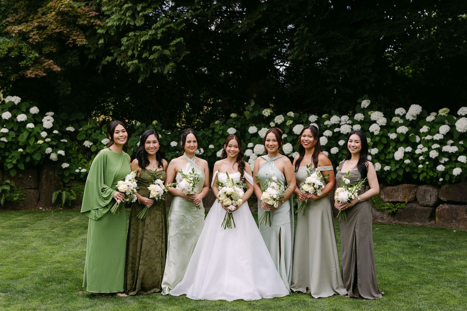 Bride in white gown poses with six bridesmaids in mismatched green dresses, holding bouquets before white hydrangeas.