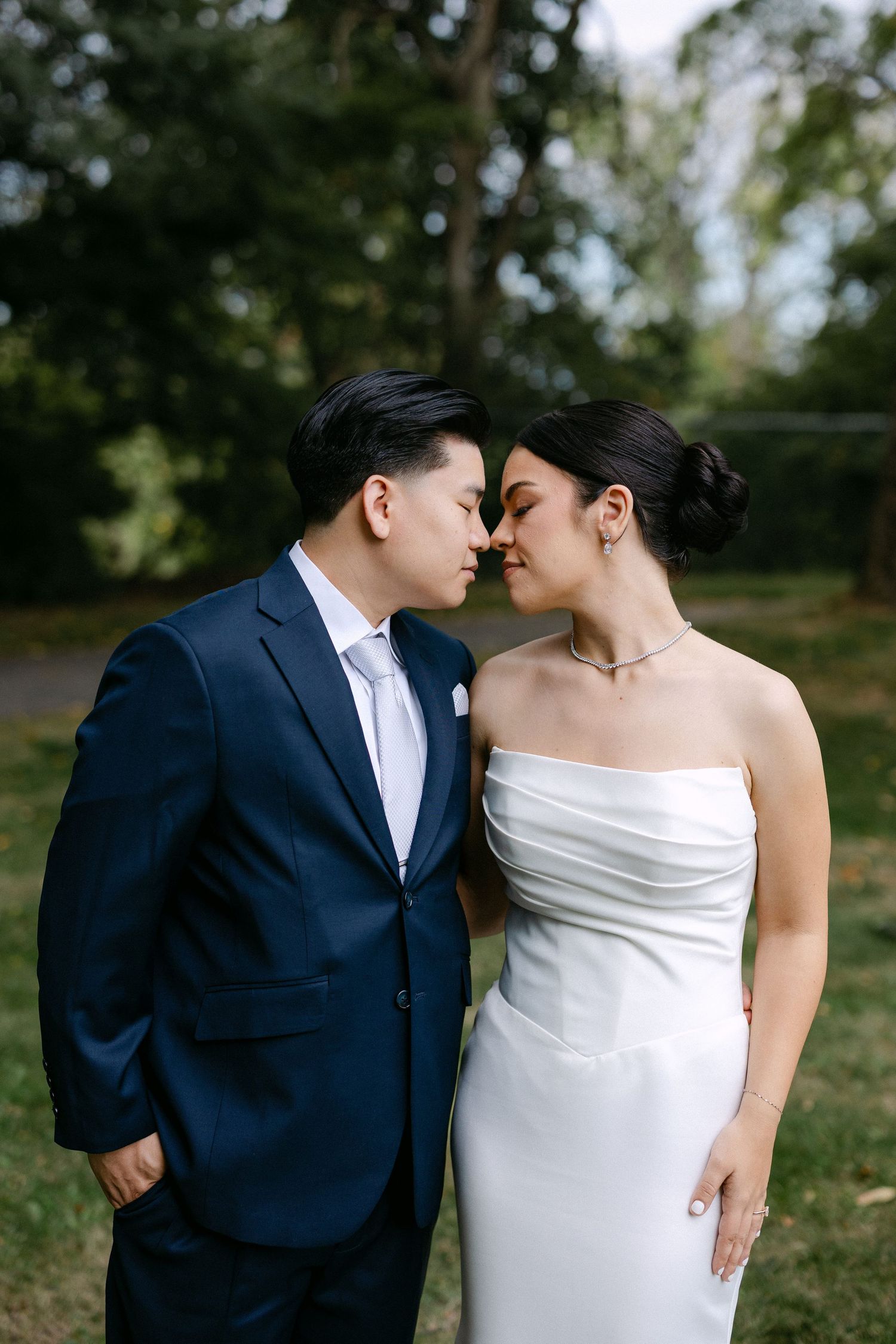 A couple in wedding attire shares a romantic nose-to-nose moment outdoors, groom in navy suit, bride in white strapless gown.