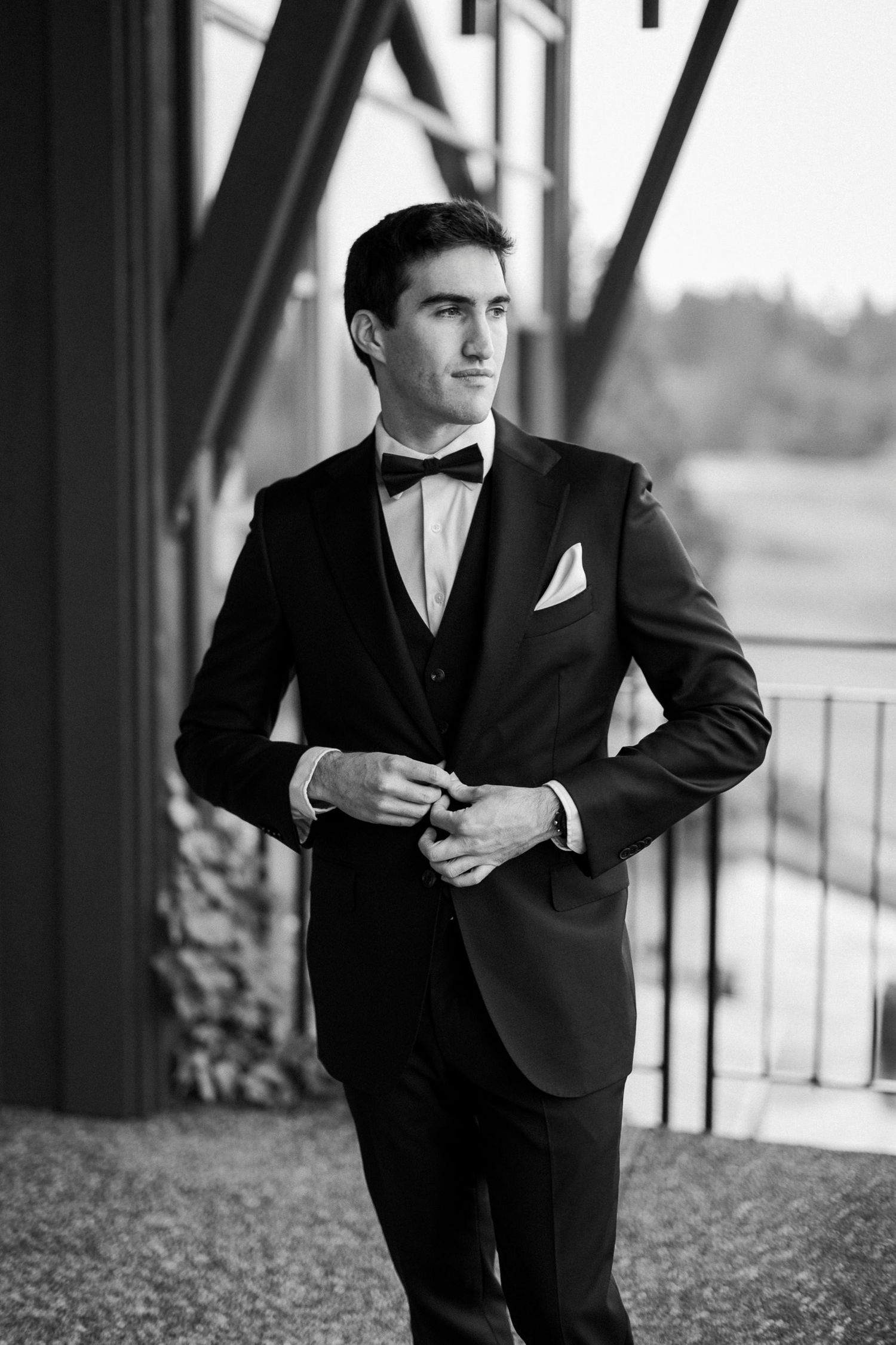 A dapper groom in a black tuxedo with bow tie adjusts his jacket outdoors in a black and white portrait.