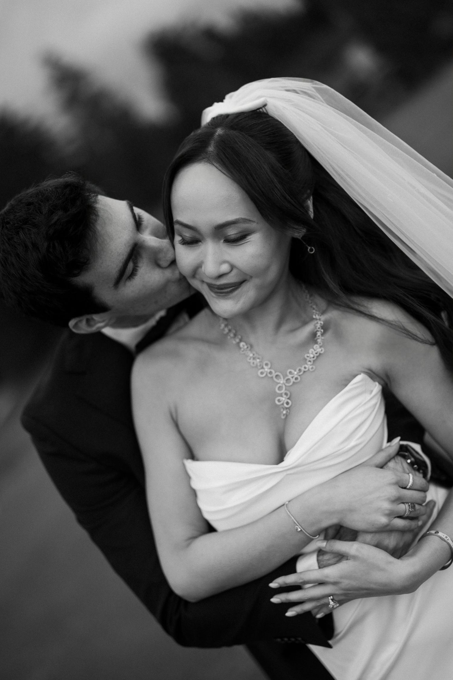Black and white wedding photo of groom kissing bride on cheek, bride smiling in strapless gown and veil.