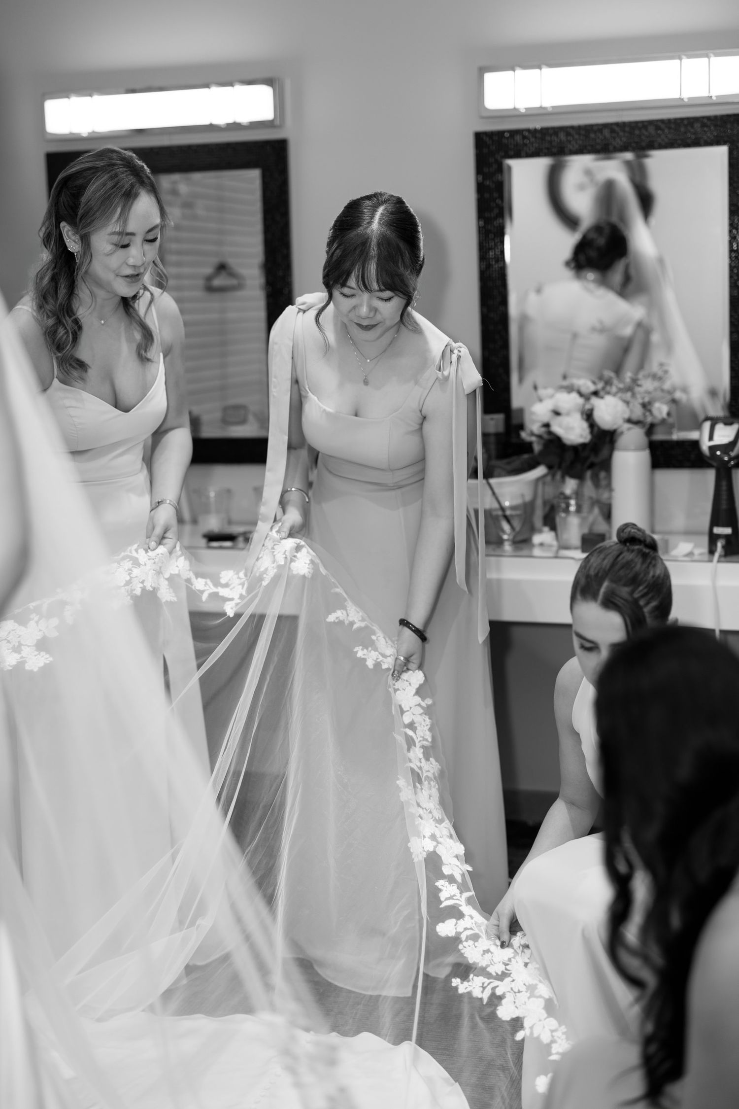Bridesmaids helping bride with floral lace veil in bridal suite, black and white wedding preparation photo.