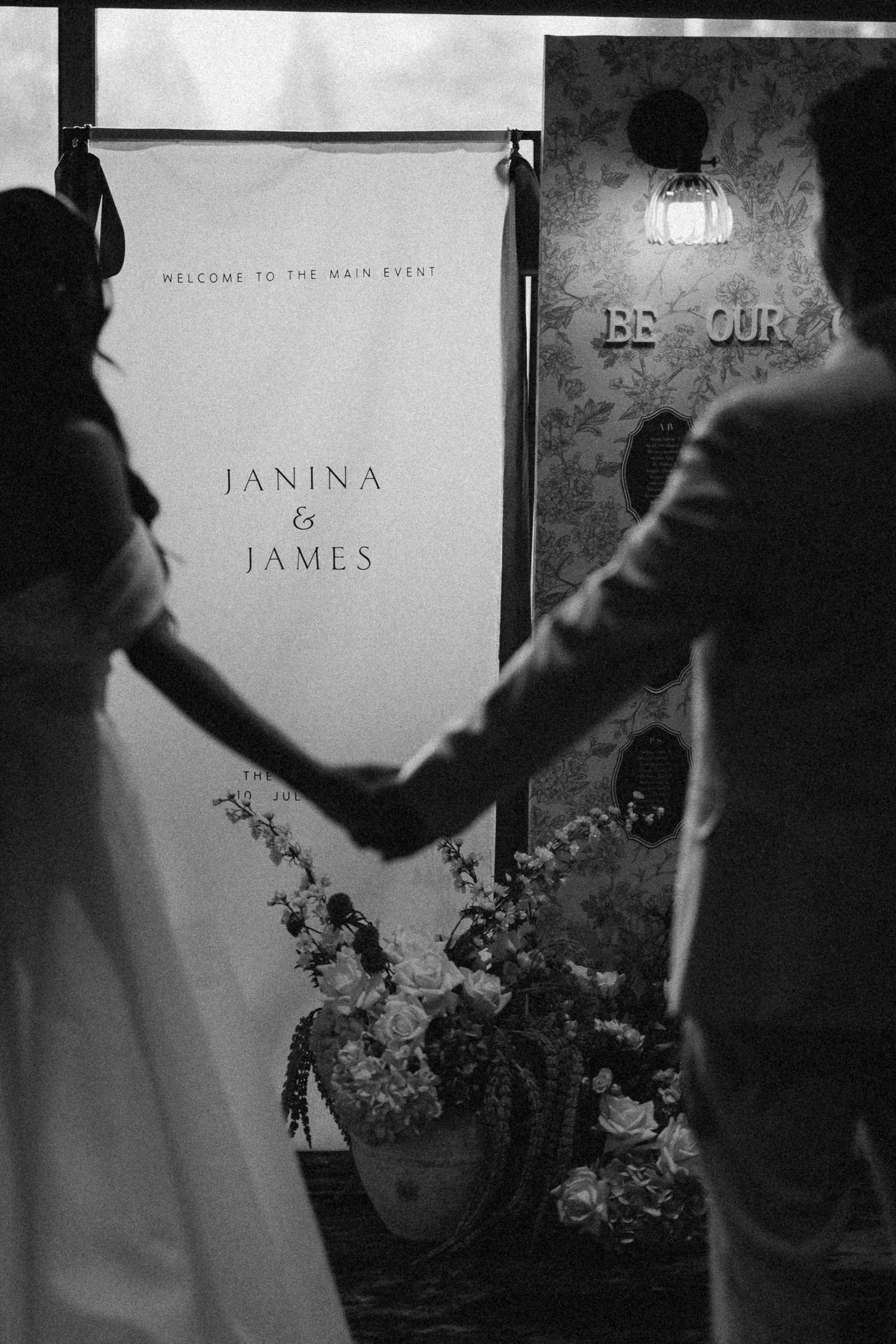 Black and white photo of Janina and James holding hands before their wedding welcome sign with floral arrangements.