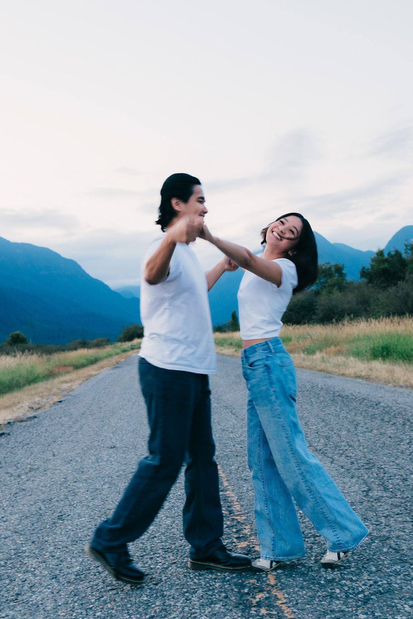 Couple in white tees and jeans dance and laugh on an empty mountain road at dusk.