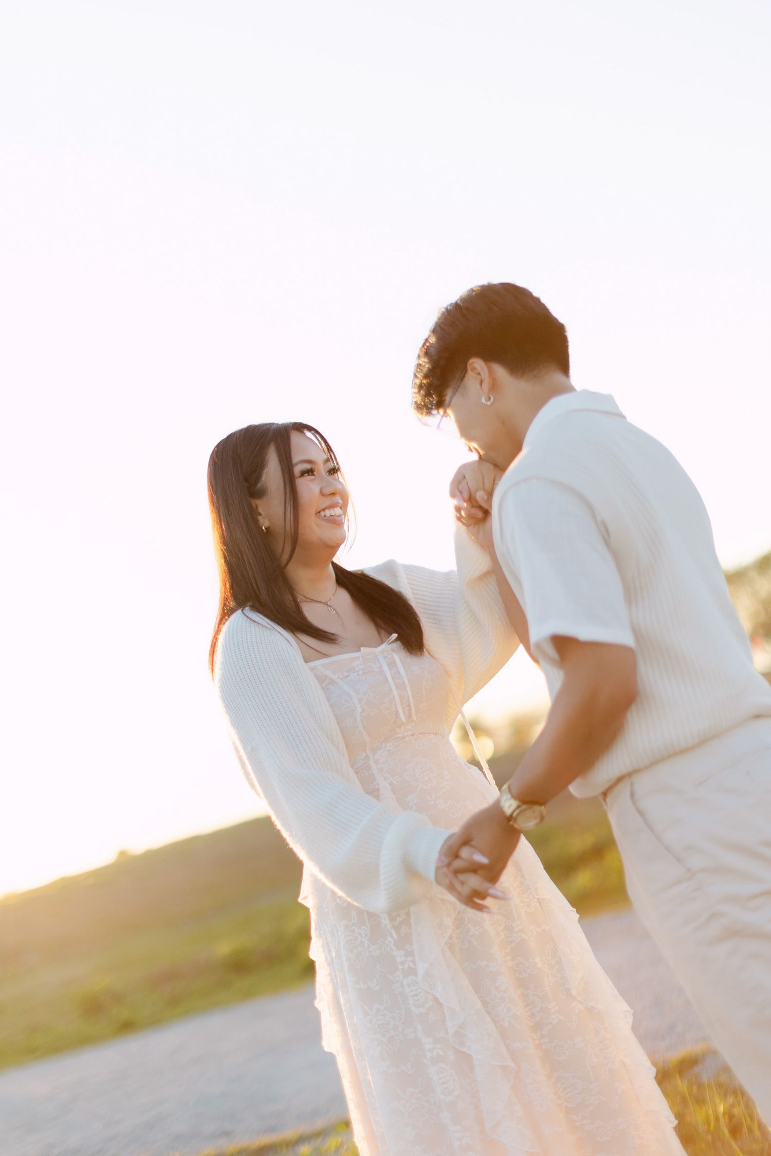 Man kissing his fiancée's hand as she smiles in a white lace dress during a sunset engagement photoshoot.