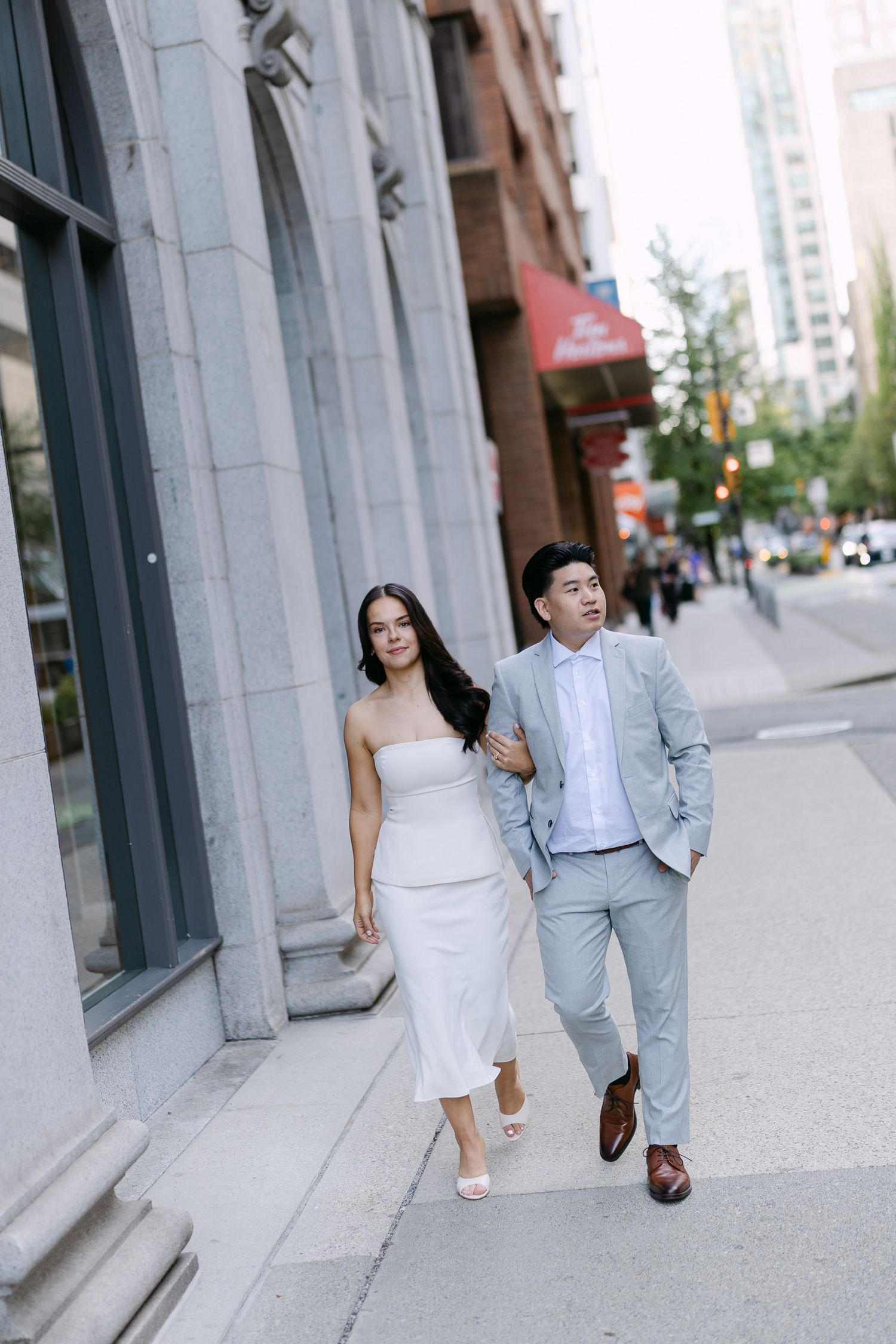 Couple walk arm in arm along a downtown city sidewalk, the woman in a white dress and the man in a light gray suit.