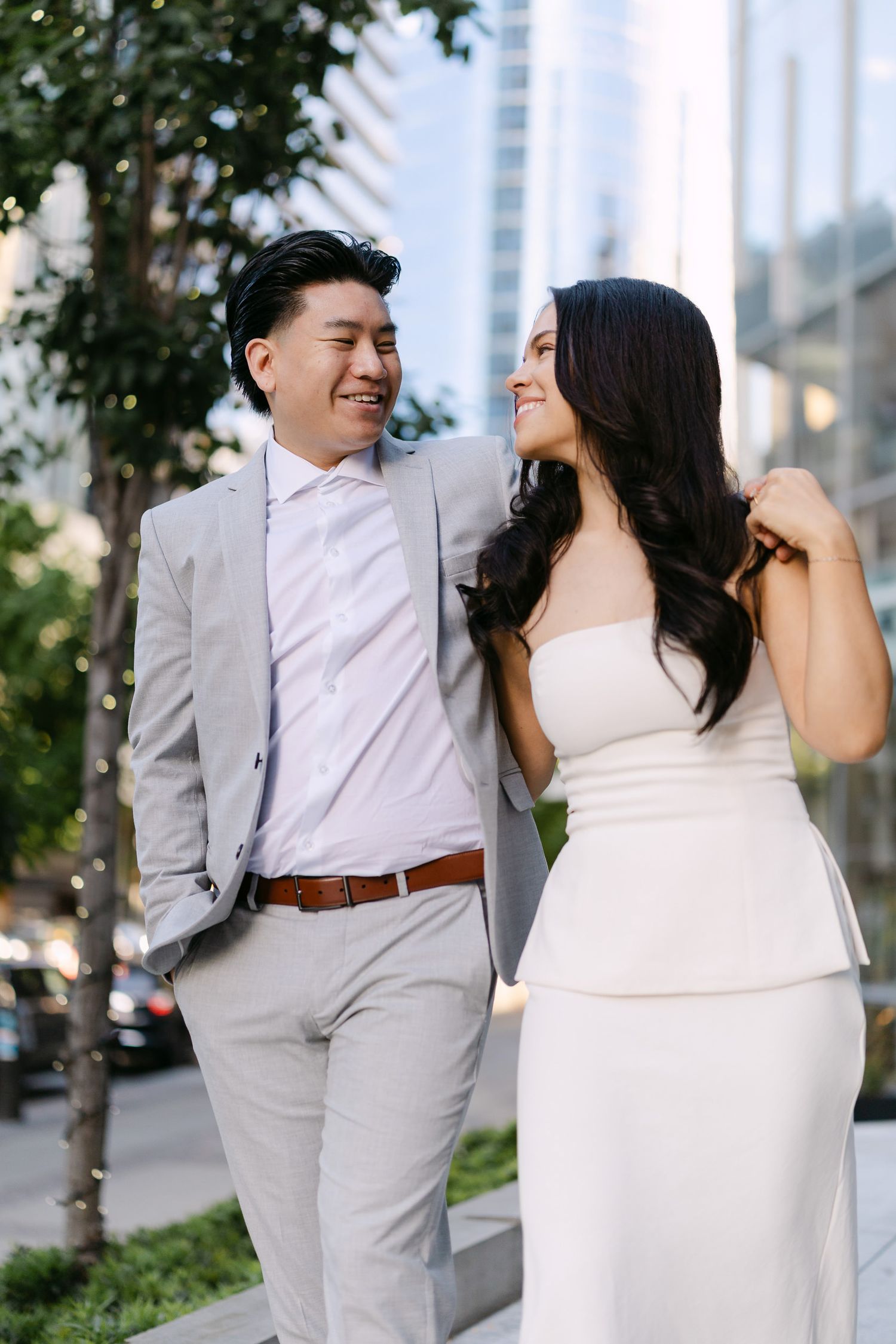Laughing couple in a gray suit and white dress share a joyful moment on a tree-lined city street during an engagement shoot.