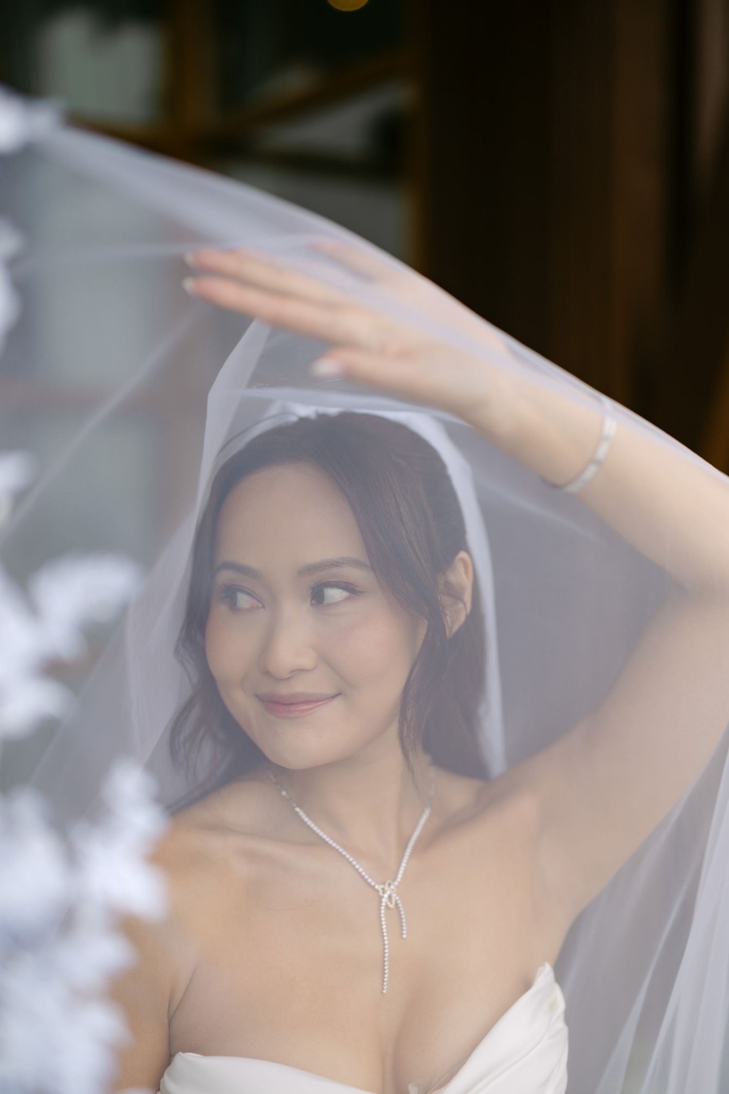 A beautiful Asian bride in a white strapless gown lifts her floral veil, wearing a delicate diamond necklace and bracelet.