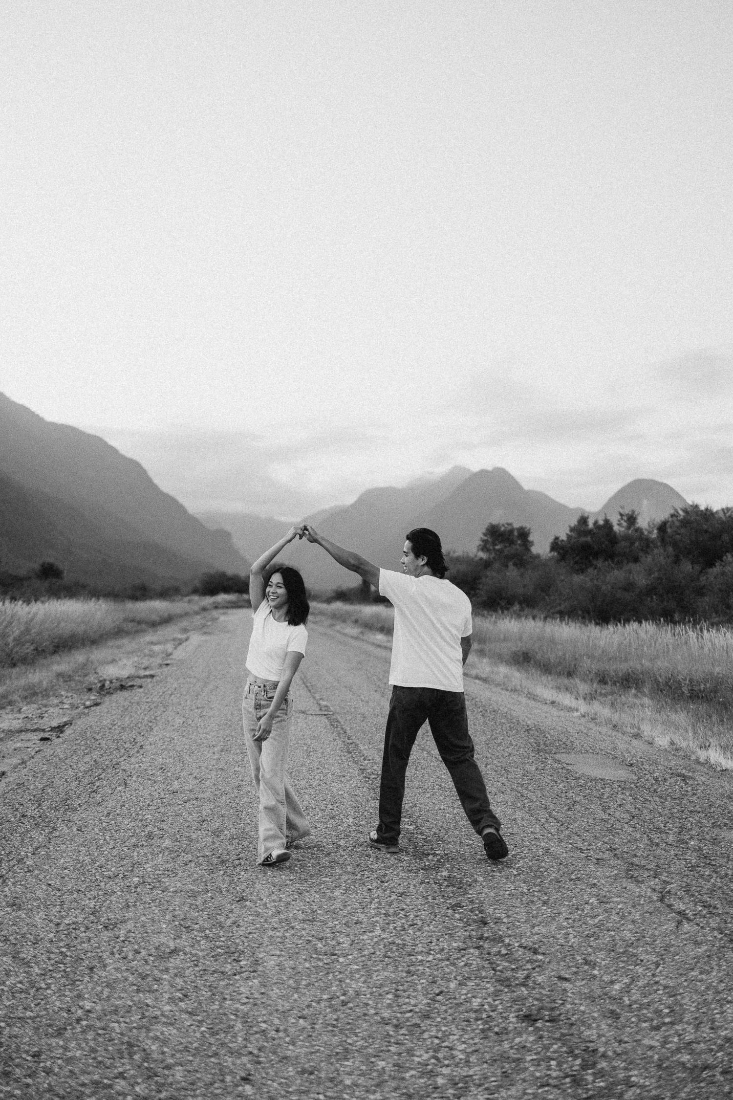 Playful black and white sequence of two people dancing and spinning on a gravel road with mountains in background.