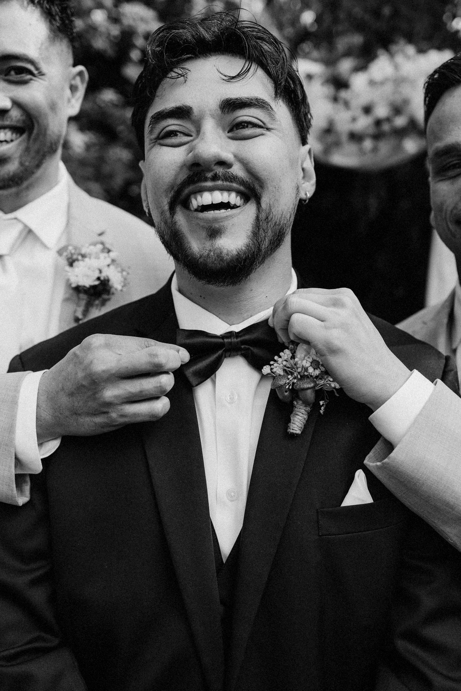 Black and white photo of a joyful moment at a wedding as a bow tie is adjusted, with boutonniere and formal attire.