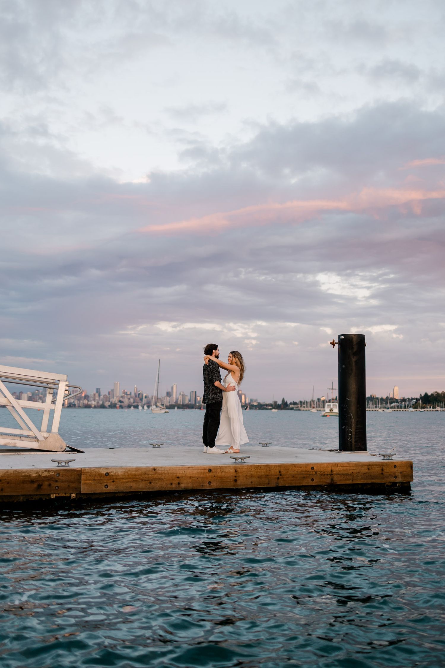 A romantic seaside photo shoot on a wooden dock at sunset with a city skyline in the background.