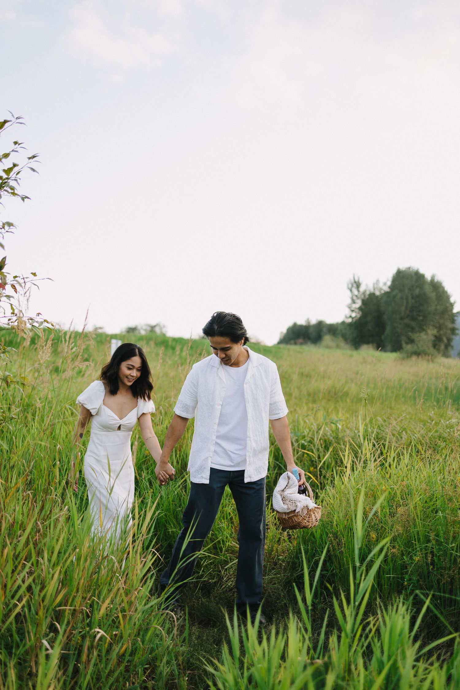 A couple in white clothing walks hand in hand through a lush green meadow on a summer day.