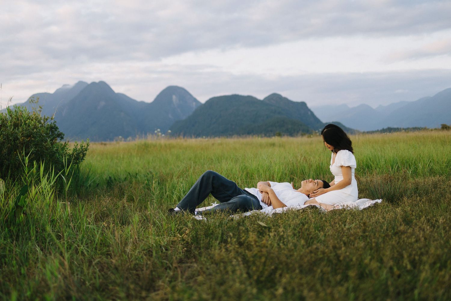 A couple relaxes on a picnic blanket in a green meadow with mountain peaks rising in the background.