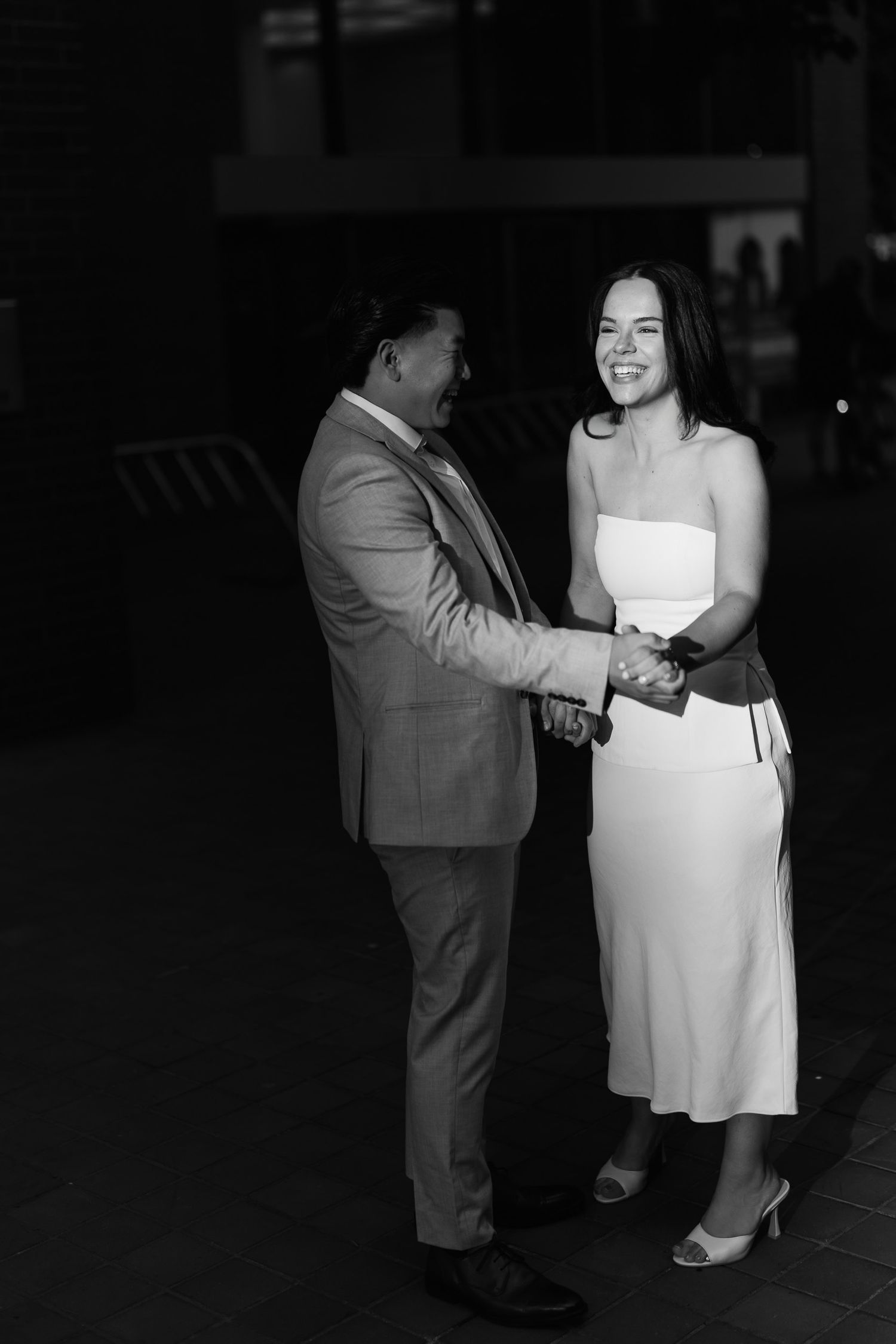 Black and white photo of a laughing couple holding hands, the woman in a white dress and the man in a suit.