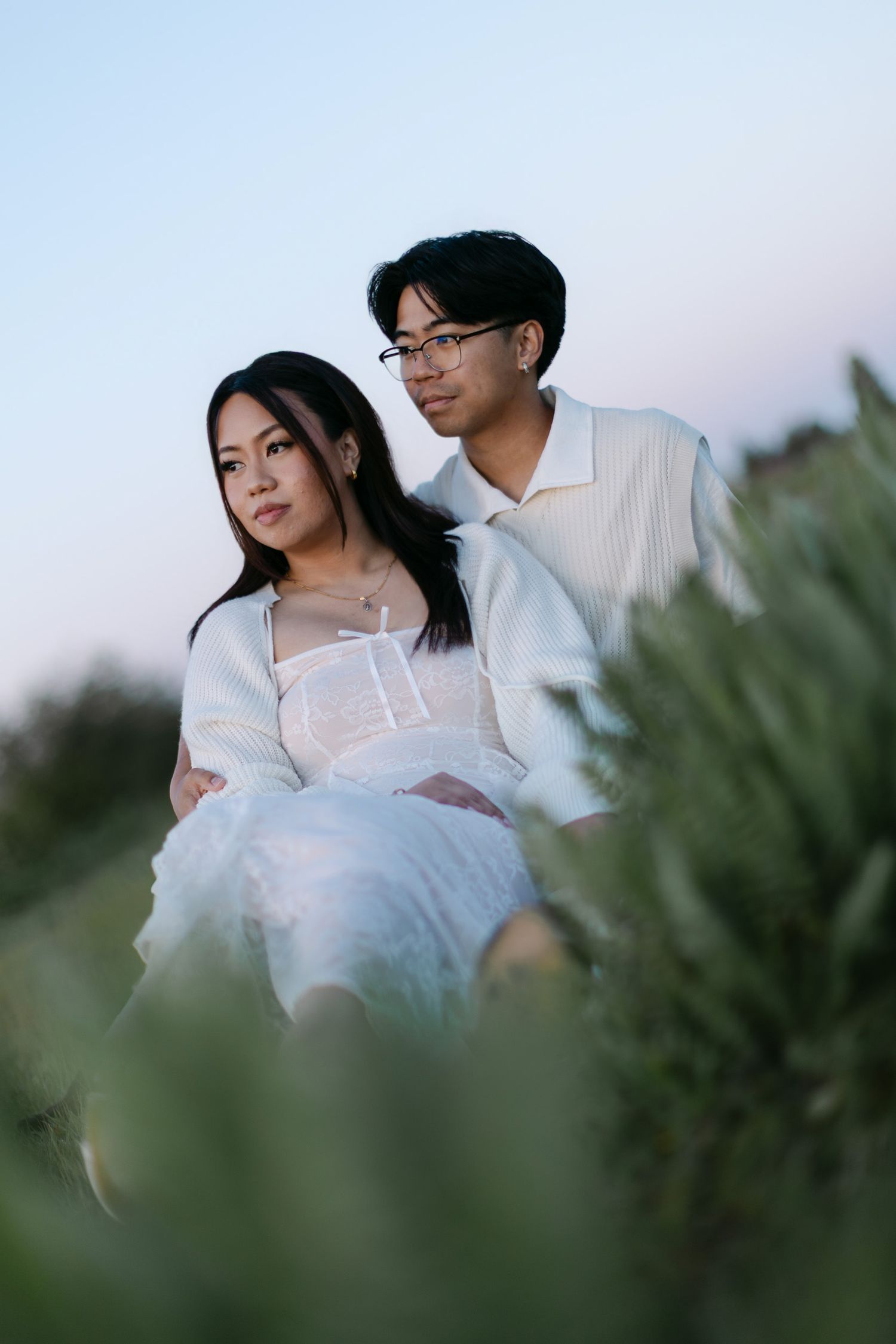 Couple in white outfits sit close together among green coastal shrubs at dusk during an outdoor engagement photo session.