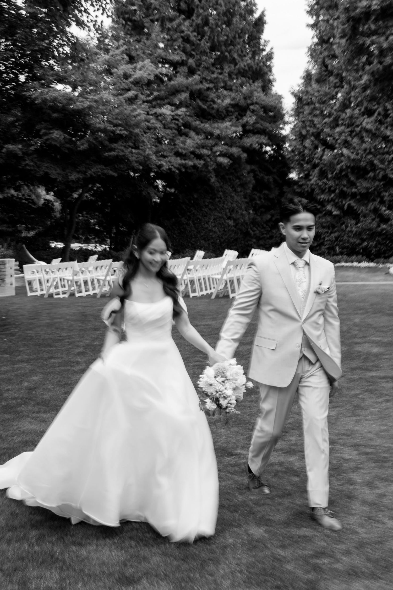Bride and groom walk hand-in-hand across a lawn after their outdoor wedding ceremony in a black and white photo.