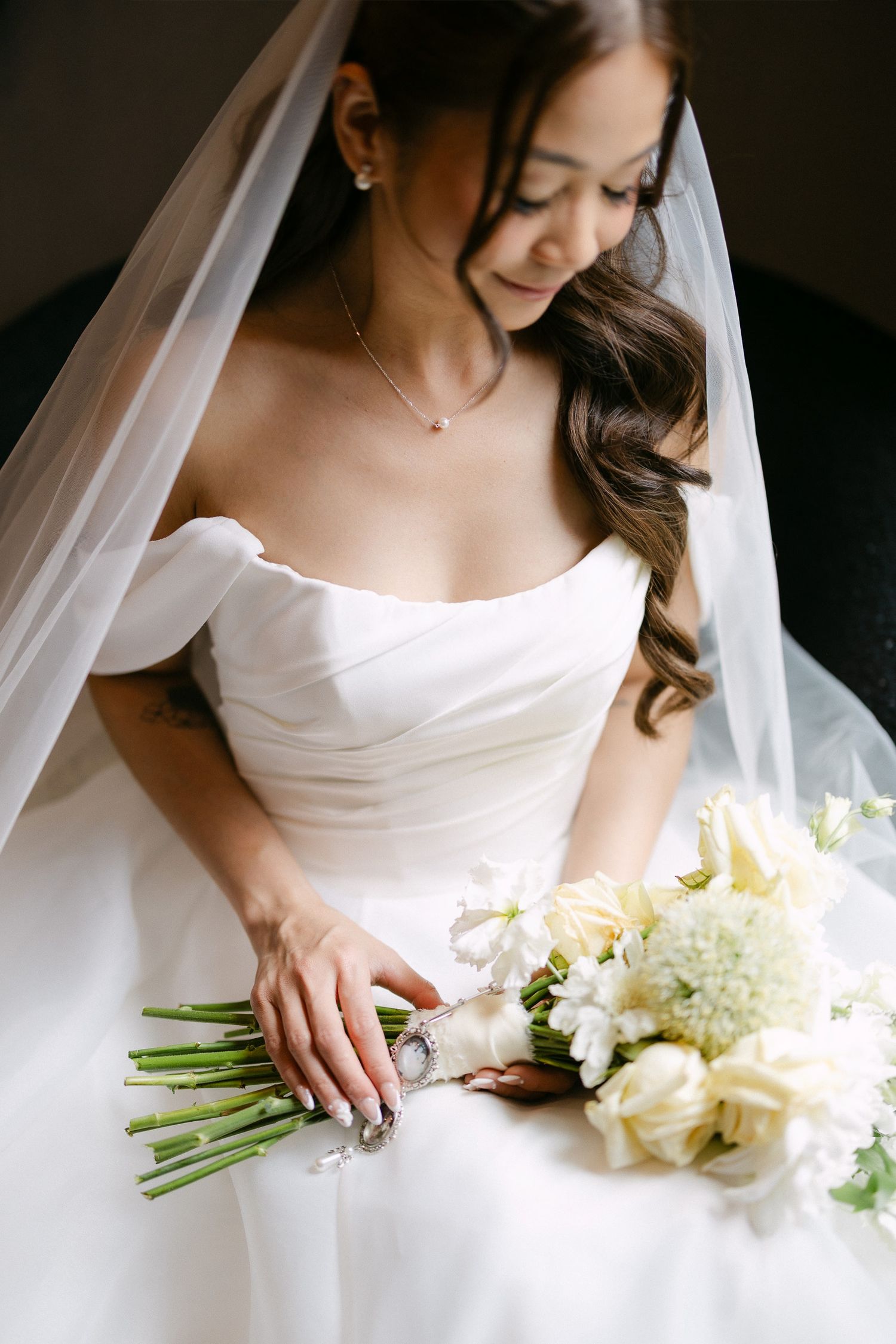 Bride in off-shoulder white gown and veil holds elegant white floral bouquet, gazing down with a serene smile.