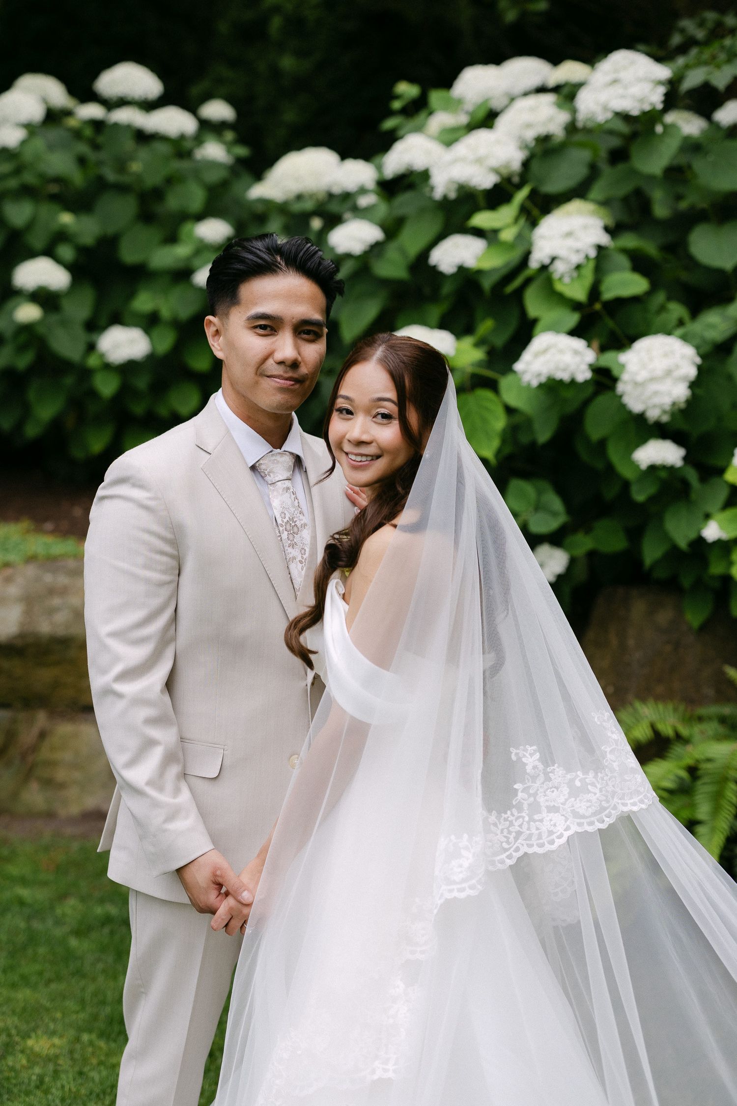 A smiling bride in a white gown and veil holds hands with a groom in a beige suit, posing before blooming hydrangeas.