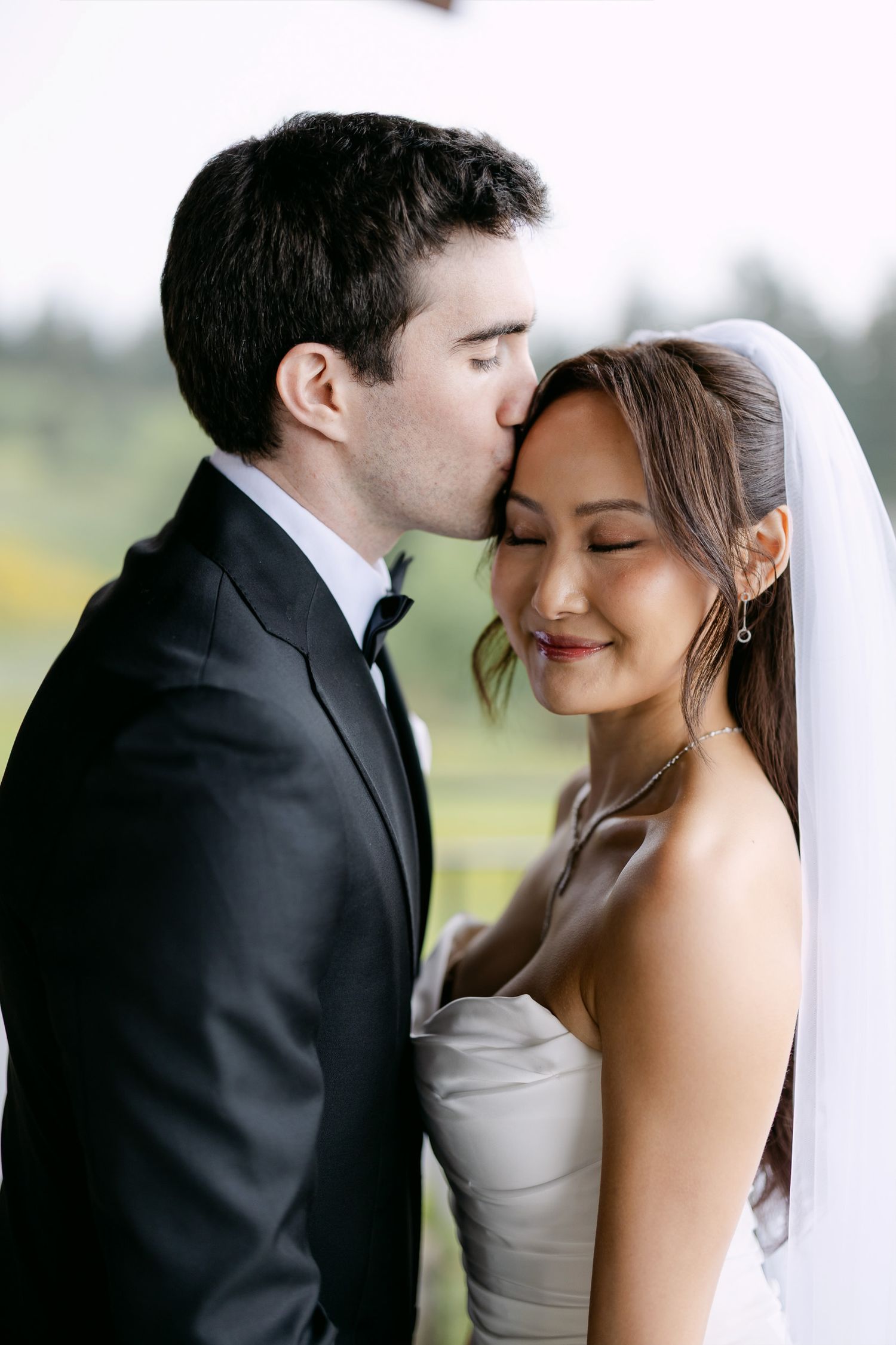 Groom in black tuxedo kissing bride on forehead as she smiles in white strapless gown and veil.