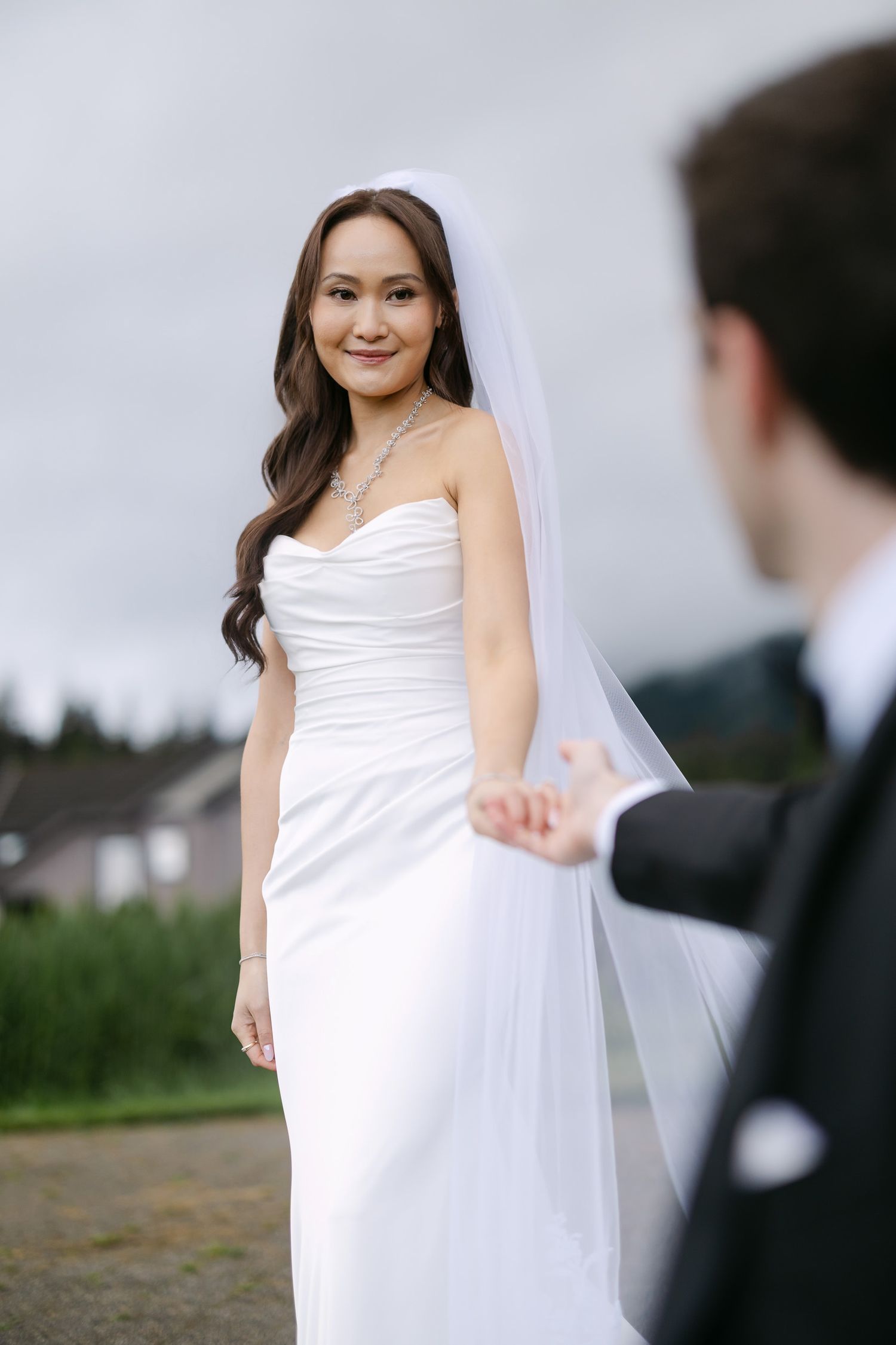 Smiling bride in elegant white ruched gown and veil holds grooms hand outdoors.