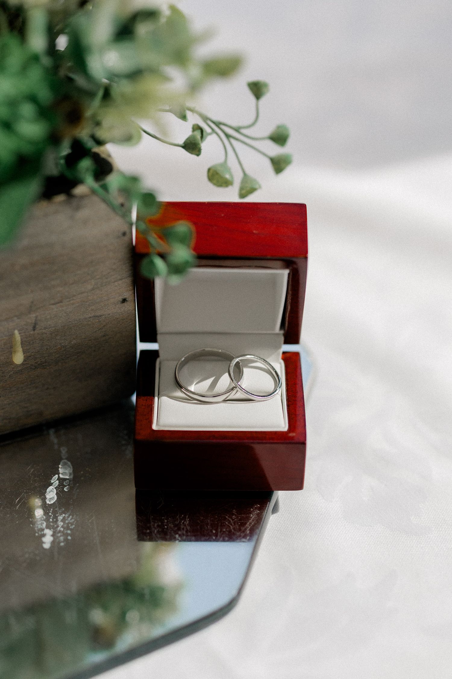 Two silver wedding rings nestled in an elegant mahogany wooden ring box with green foliage decor.