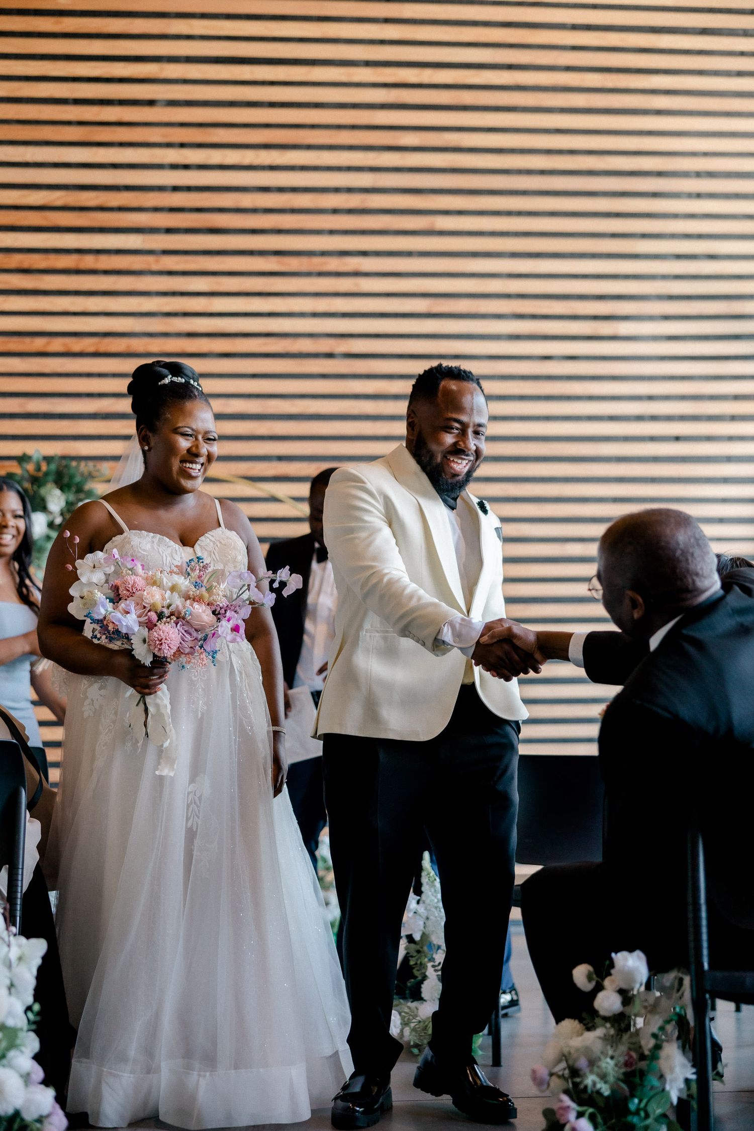 A joyful wedding ceremony takes place in front of a wooden slatted wall with the couple and officiant sharing a happy moment.