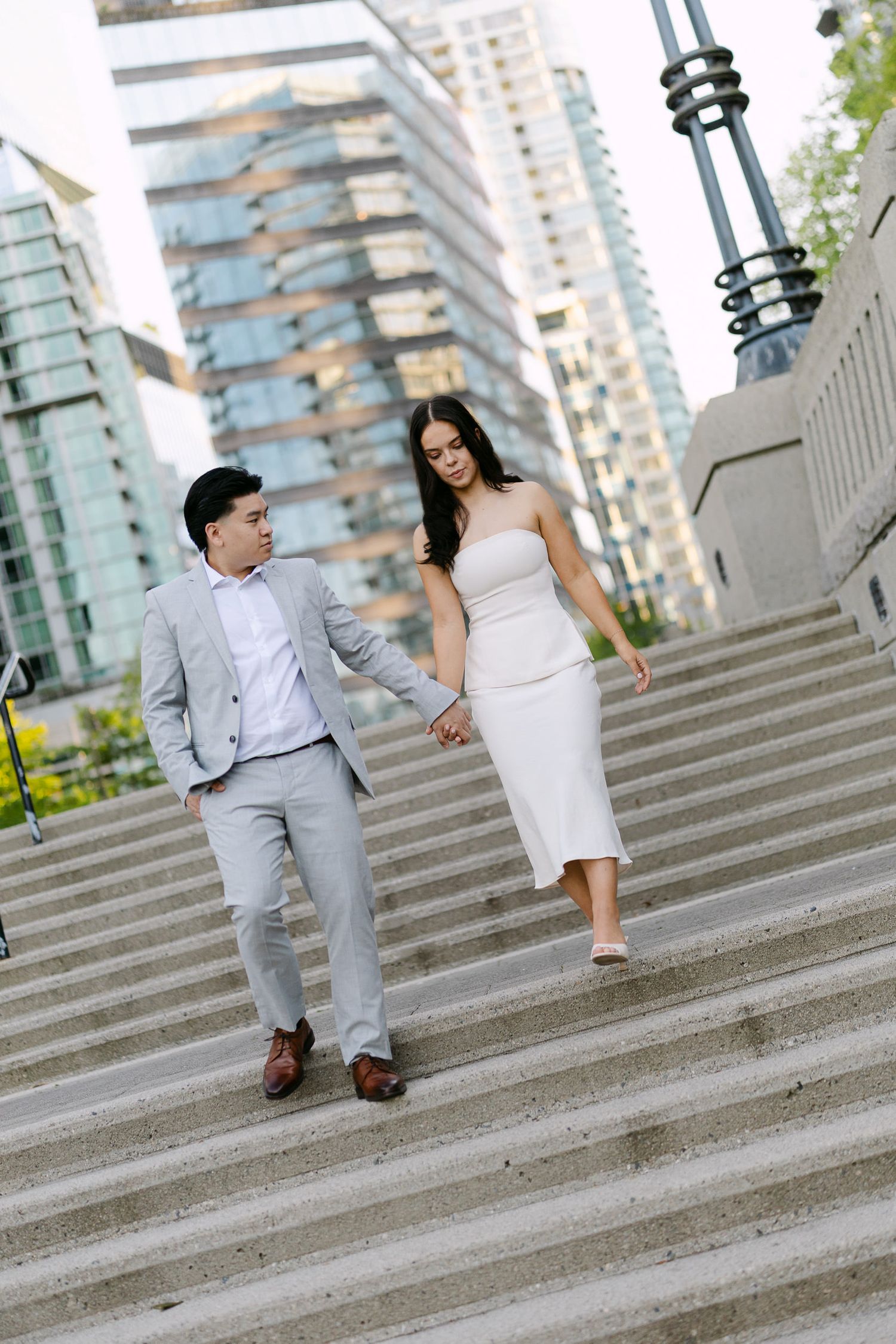 Couple in light suit and white dress holding hands while walking down city steps beneath tall glass skyscrapers.